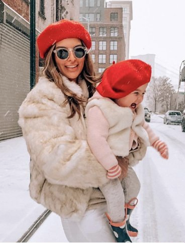Dikla laughing in the snow with her toddler daughter, dressed in matching faux fur coats and red berets.