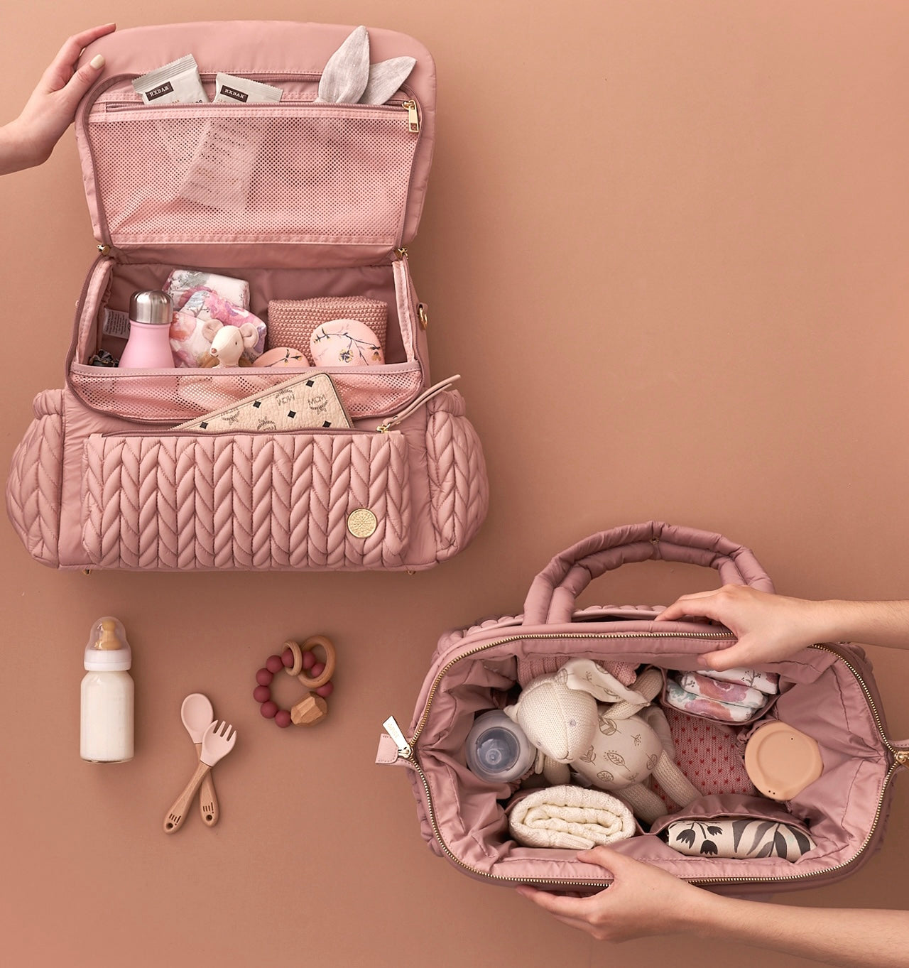 Flat lay of open Levy Backpack and Paige Carryall, both in Dusty Rose, packed with matching baby items, with hands demonstrating the wide openings of each bag.