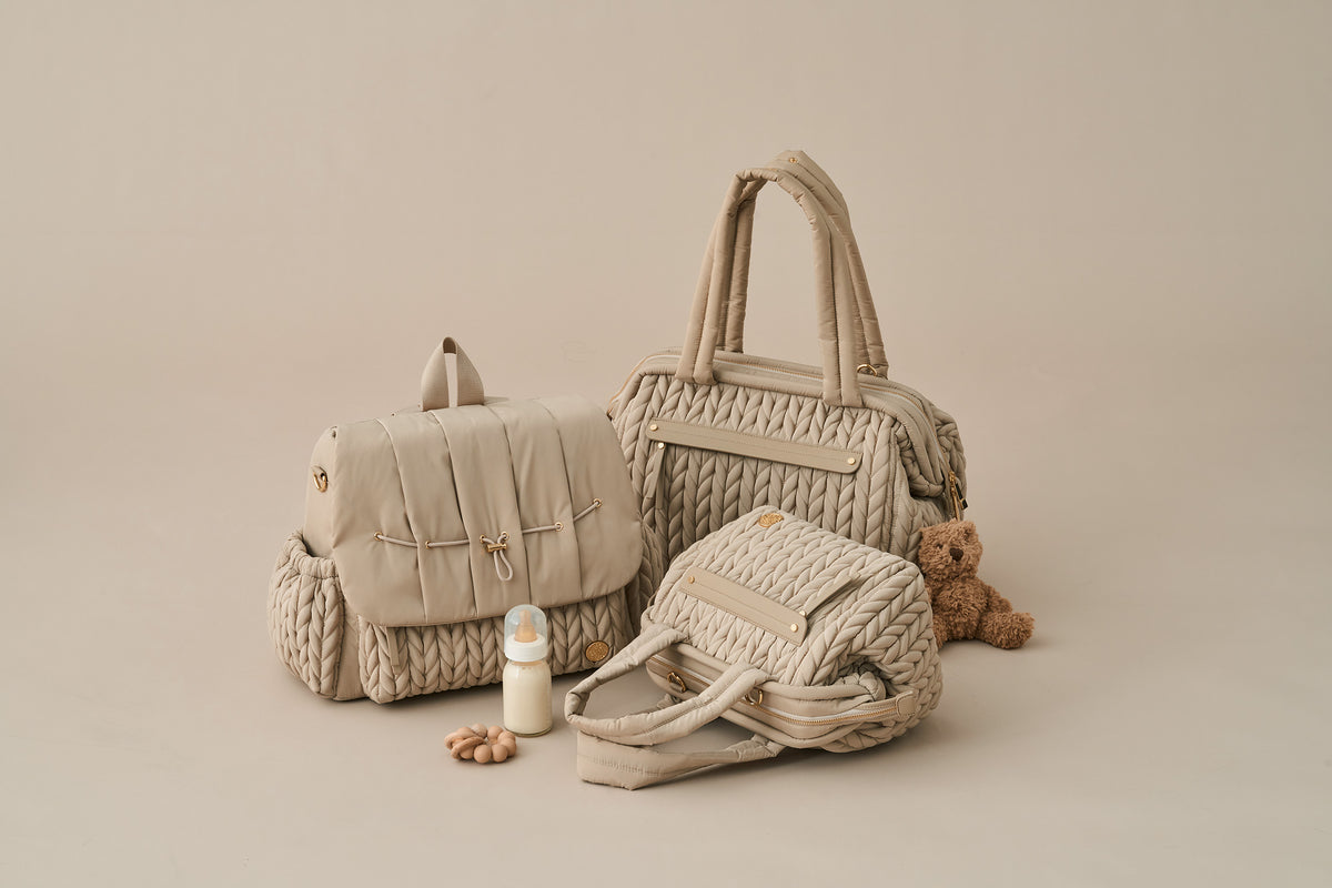 HAPP Brand Beige Collection featuring Levy Backpack and Paige Carryall standing upright, Paige Mini lying on its side, with a brown teddy bear, teether, and milk bottle on a beige background.