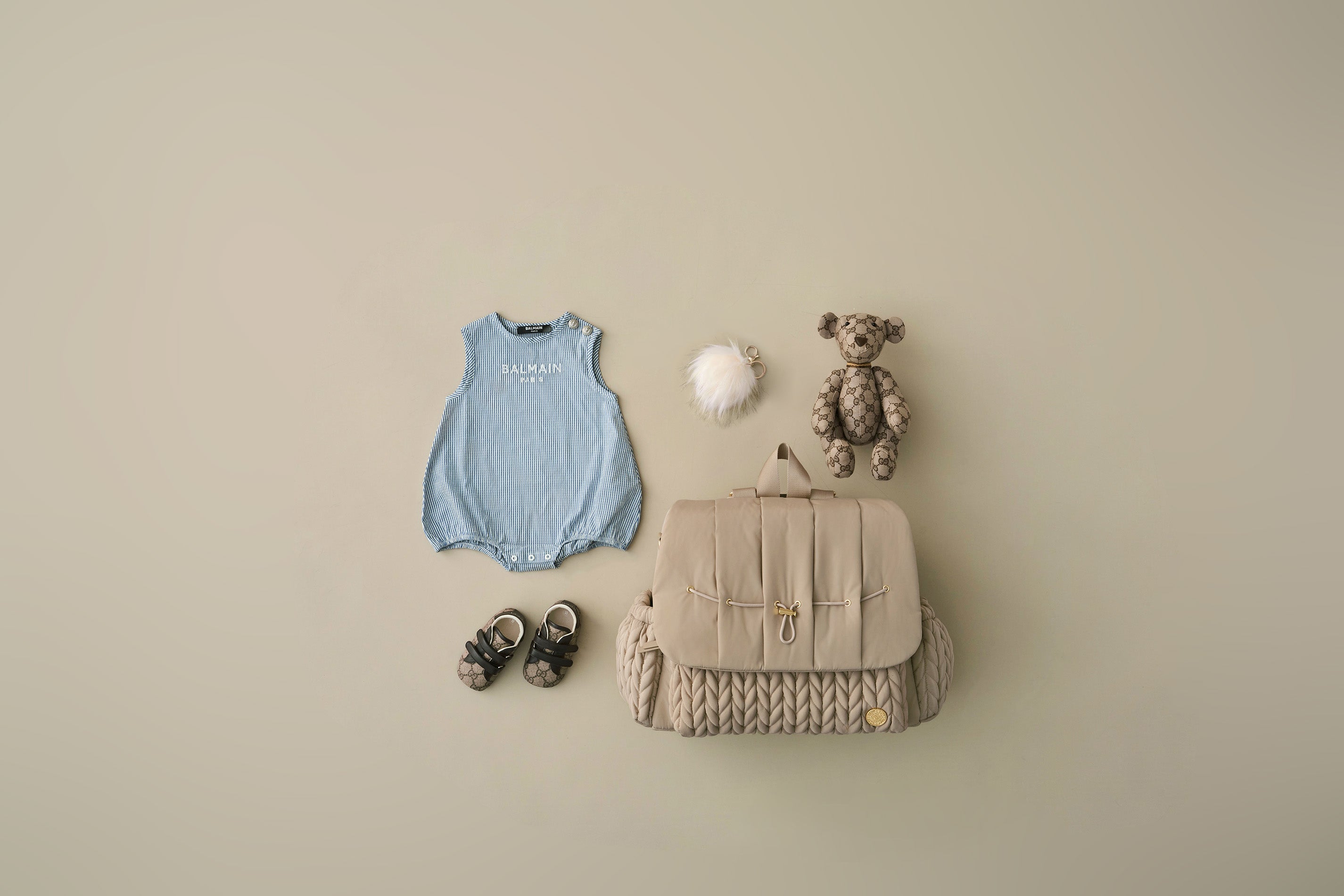 HAPP Brand Levy Backpack shown closed beside Pouf Charm free gift, alongside matching blue and brown baby items; shoes, a teddy bear, and a onesie. Flat lay on a beige background.