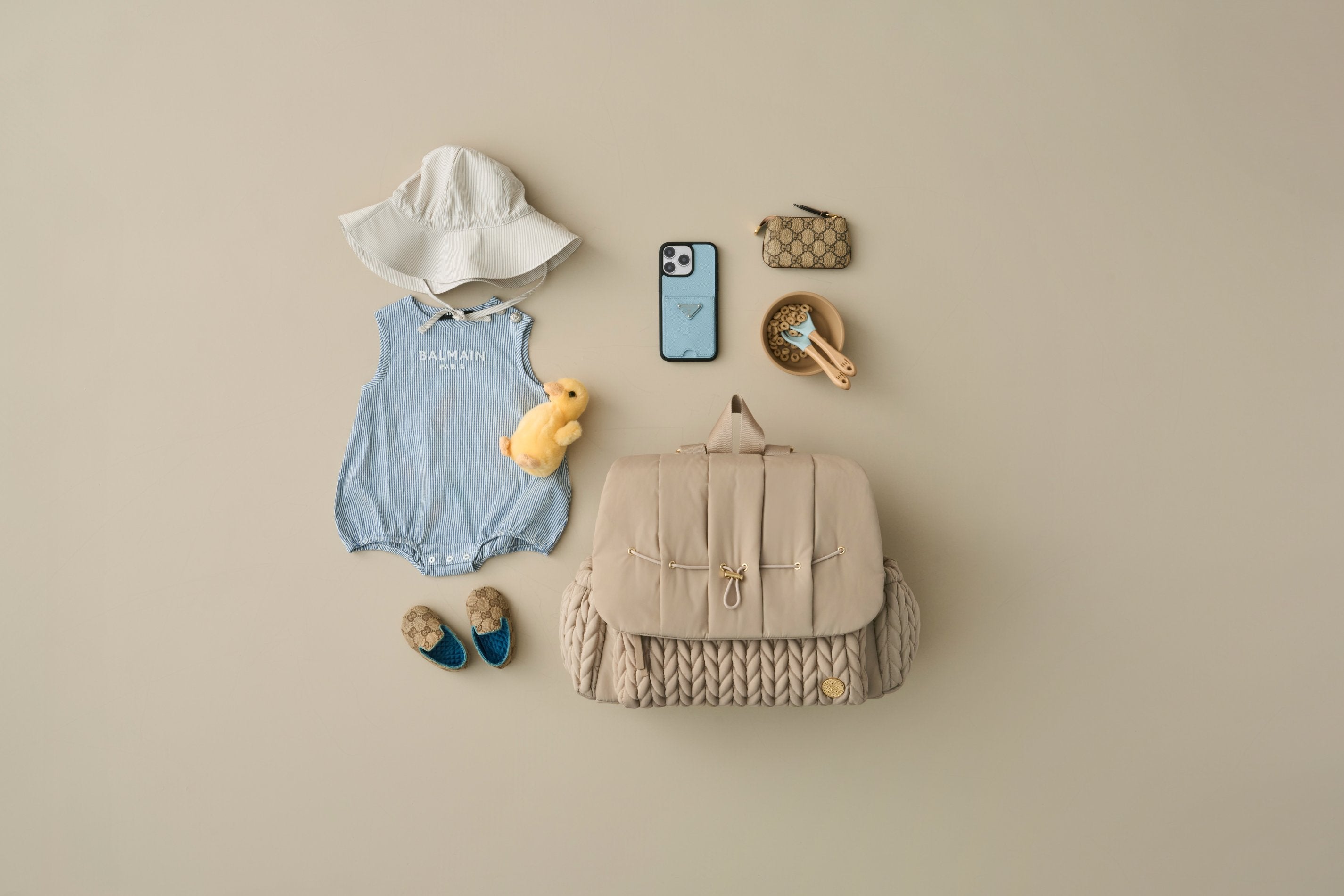 HAPP Brand Levy Backpack in Beige pictured with blue onesie, white baby sunhat, beige baby shoes, yellow stuffed duckie, phone in blue case, beige wallet, and a bowl of baby cereal with blue baby utensils on a beige background.