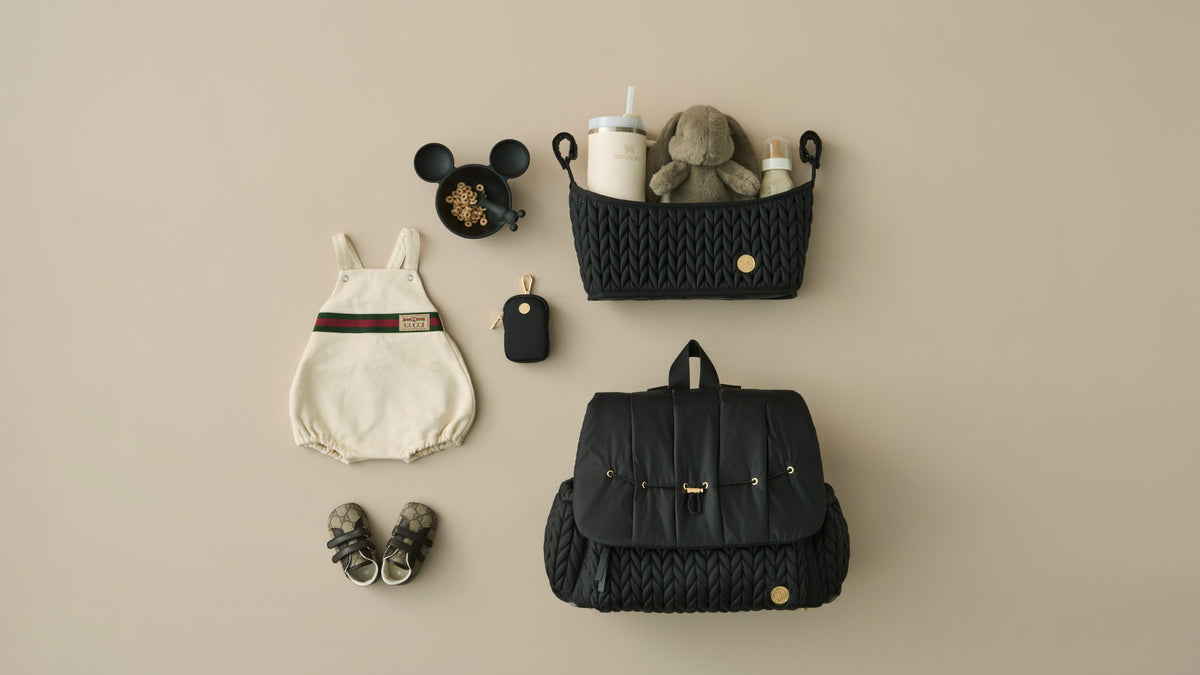 Flat lay image featuring HAPP Brand Levy Backpack, Stroller Organizer, and Clip-on Pouch in Classic Black, arranged with beige and brown baby items on a beige background.