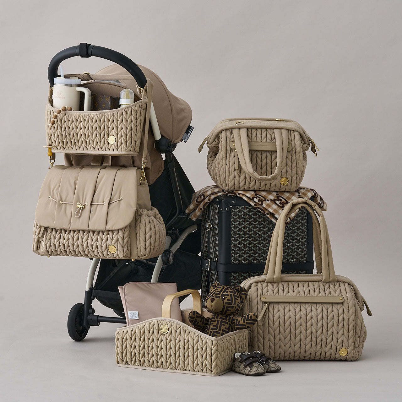 The HAPP Brand Beige collection all together, including Levy Backpack, Stroller Organizer, Paige Mini, Paige Carryall, and Diaper Caddy, arranged on and around a stroller and suitcase.
