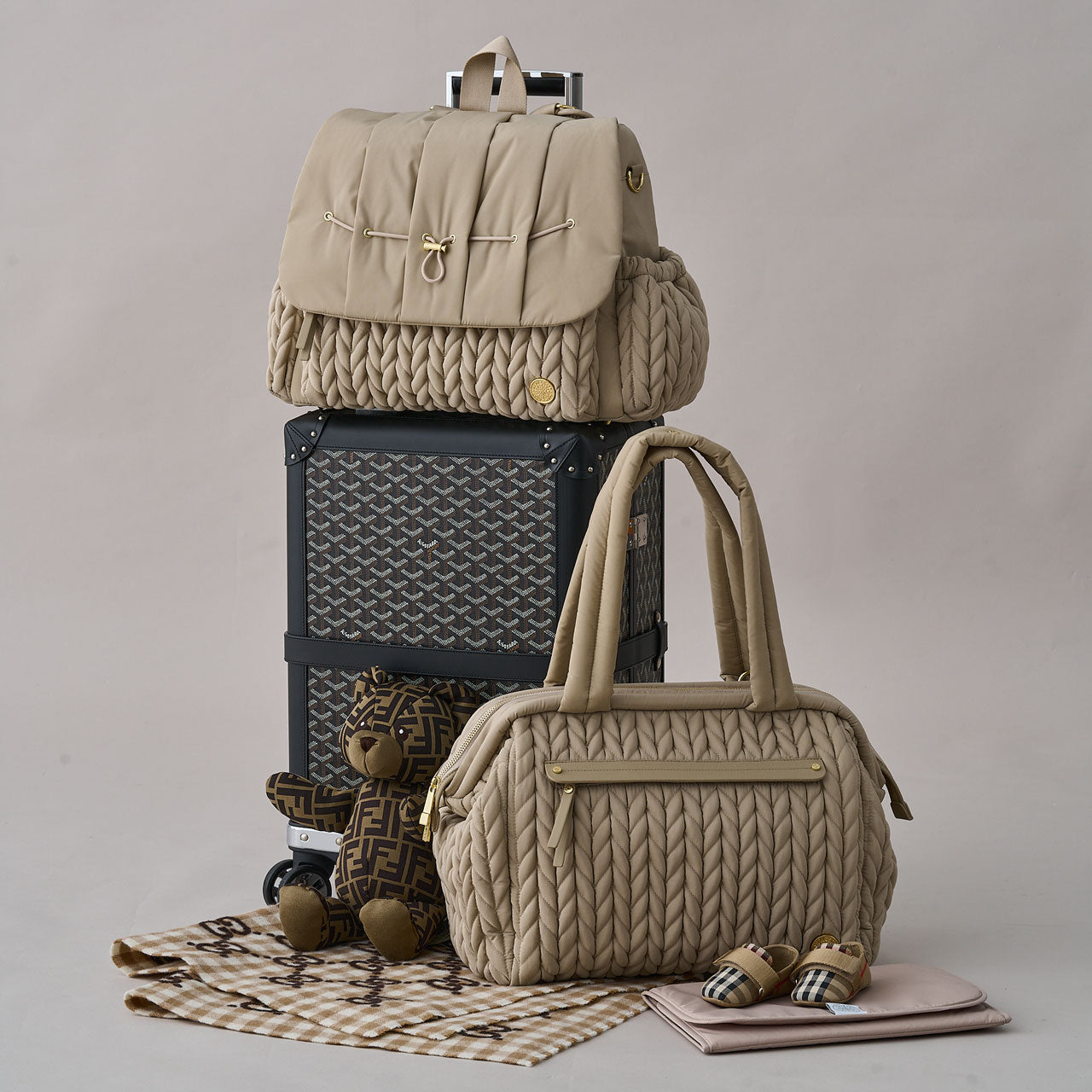 Levy Backpack Beige sitting on top of a black patterned rolling suitcase, with the Paige Carryall Beige sitting beside it atop a beige blanket next to a teddy bear.