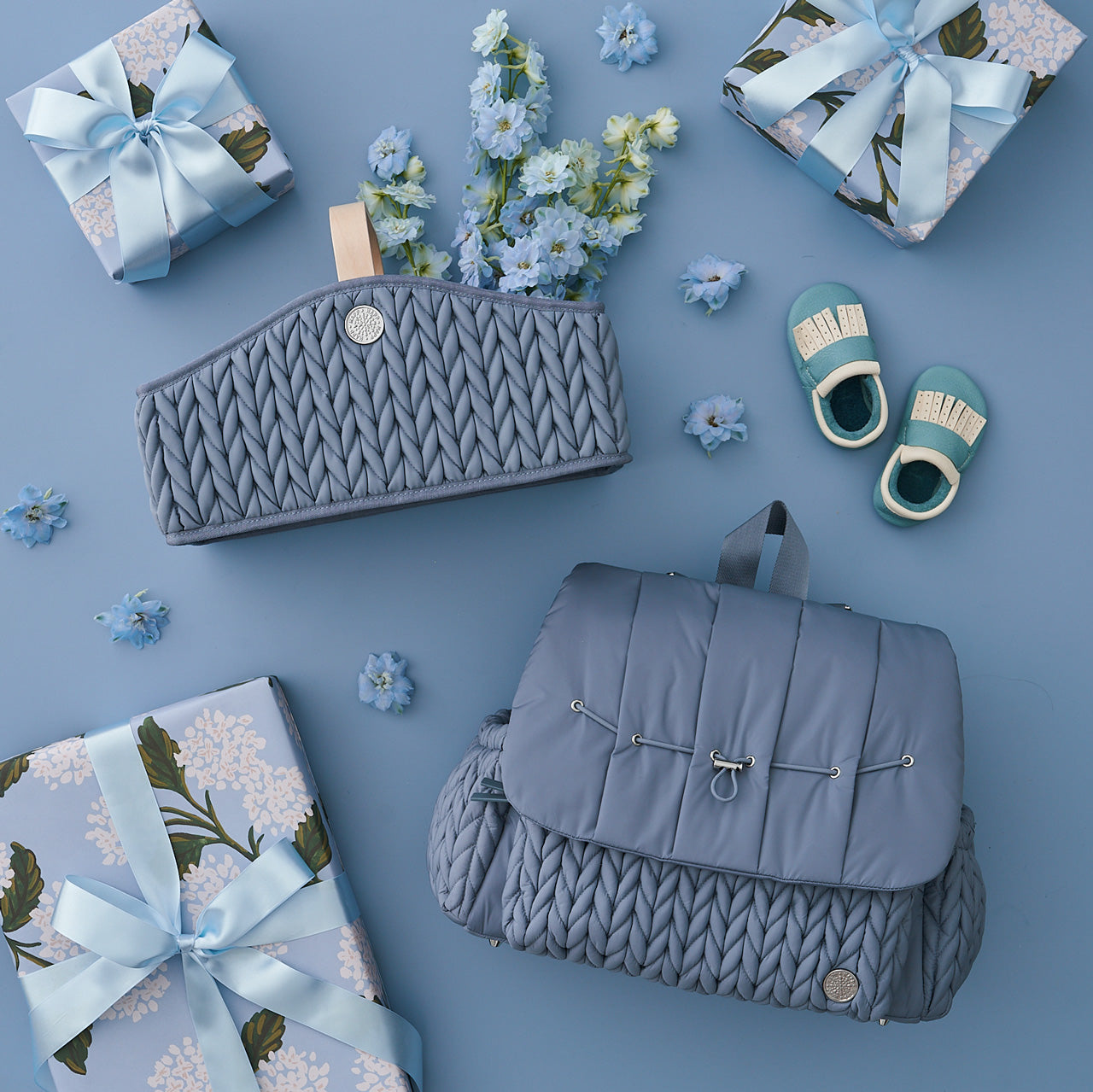 Flat lay with HAPP Levy Backpack and Diaper Caddy in matching blue, on a blue background with matching blue baby items, shown with three wrapped gifts and blue flowers.