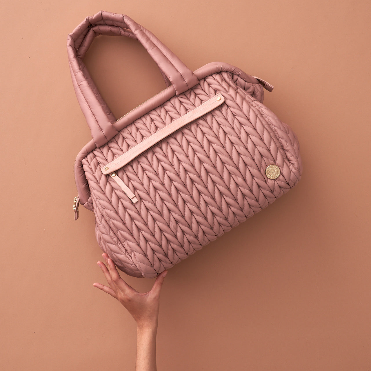 Paige Carryall Rose from the front, tilted sideways with a hand propping it up to show the size and demonstrate the light weight of the diaper bag.