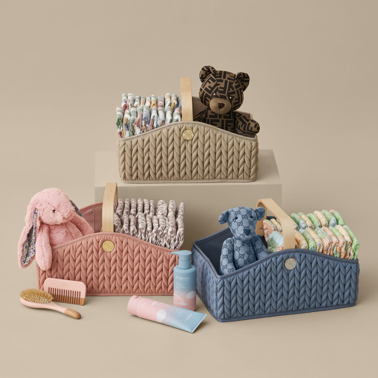 3 diaper caddies, one beige, one rose, one blue, shown standing upright and packed with diapers and matching teddy bears, arranged on blocks with a beige background.