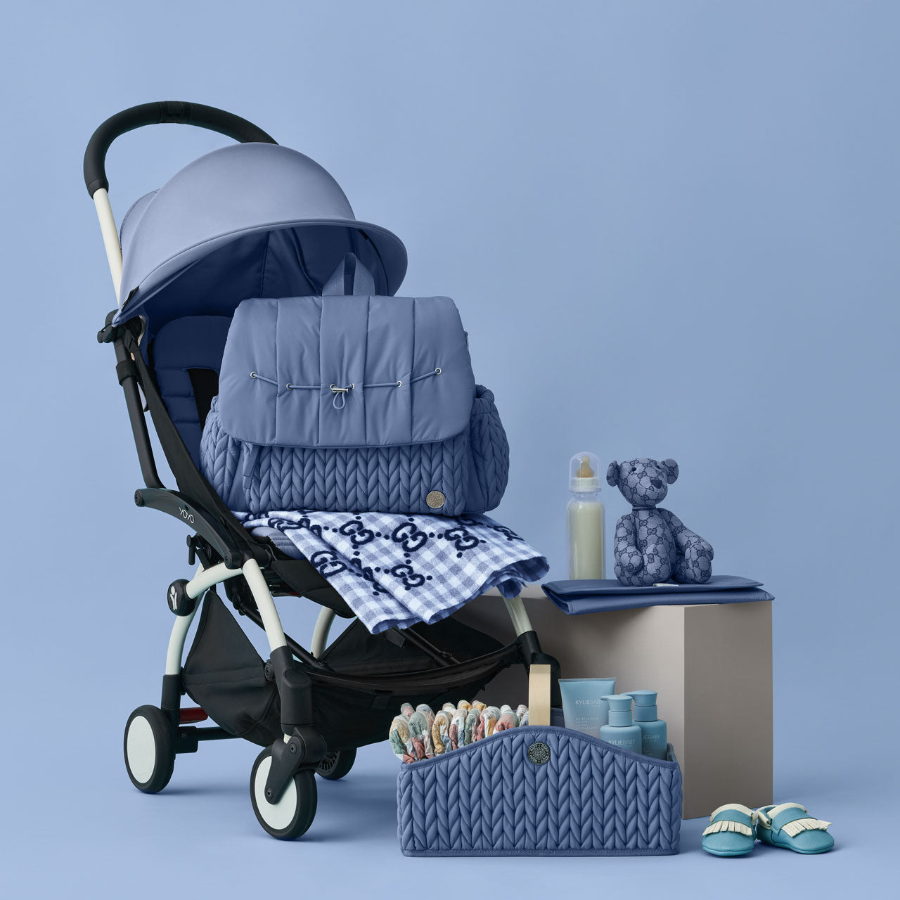 HAPP Levy Backpack and Diaper Caddy in matching blue, on a blue background, arranged on and around a blue and black stroller with matching blue baby and diapering items.