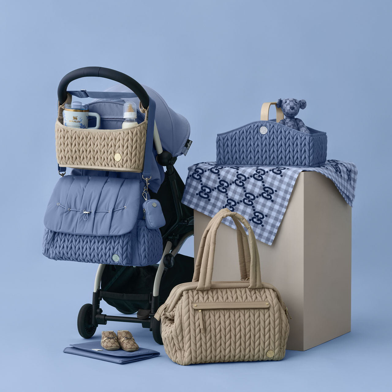 HAPP Levy Backpack in Ash Blue with Blue Clip-on Pouch attached and Stroller Organizer in Beige on a blue and black stroller; Paige Carryall Beige sitting beside it, and Diaper Caddy Blue on na block, packed with teddy bear on a blue blanket.