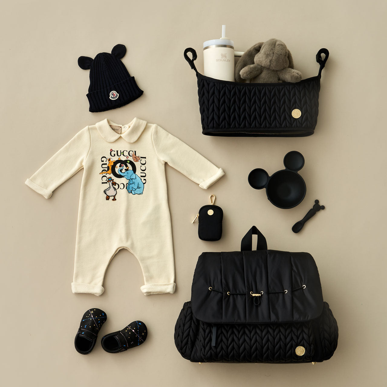 Levy Backpack in Black lying flat, showing the luxe quilted fabric, gold hardware details, convenient exterior bottle pockets, and top carry handle. Arranged with HAPP clip-on pouch in Black and black baby items on a beige background.