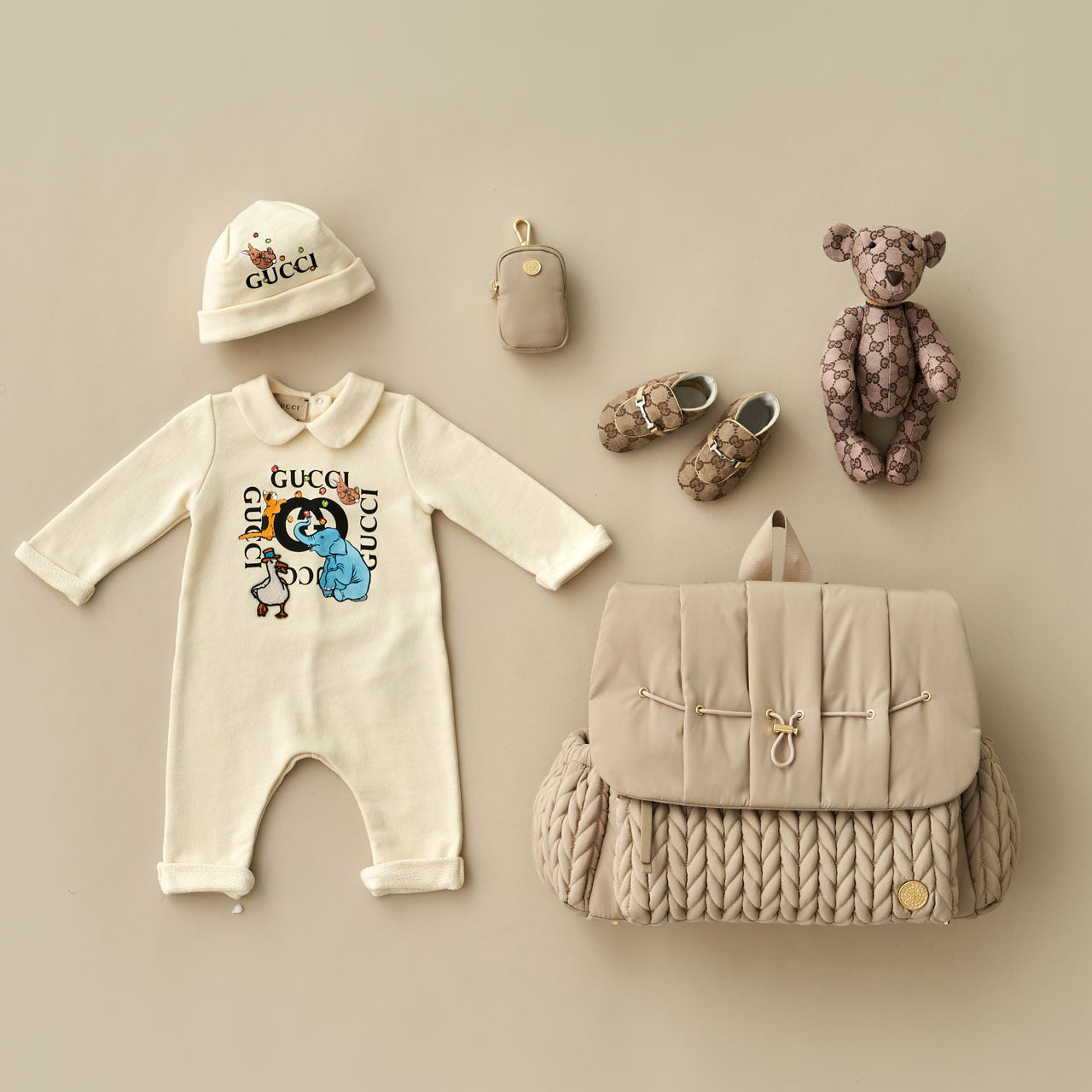 Levy Backpack in Beige lying flat, showing the luxe quilted fabric, gold hardware details, convenient exterior bottle pockets, and top carry handle. Arranged with APP Clip-on Pouch in Beige and beige baby items on a beige background.