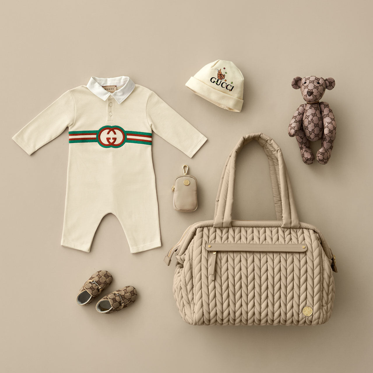 Paige Carryall Beige from the front, showing front mommy pocket, herringbone quilting, gold hardware, and padded carry handles; laid flat with matching beige and baby items and a HAPP Clip-on Pouch in Beige.