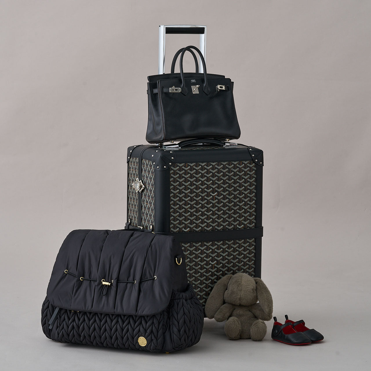Levy Backpack Black sitting beside a black patterned rolling suitcase, with a designer handbag on top and bunny rabbit teddy beside it.
