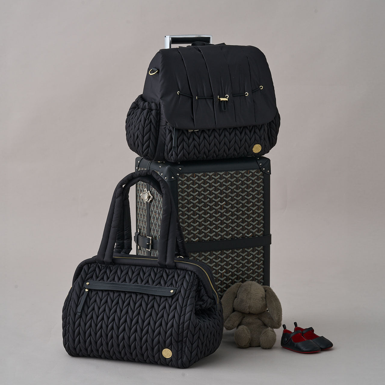 Paige Carryall Black shown with Levy Backpack Black, both on an angle showing the sides of the bags, arranged on and beside a rolling suitcase.