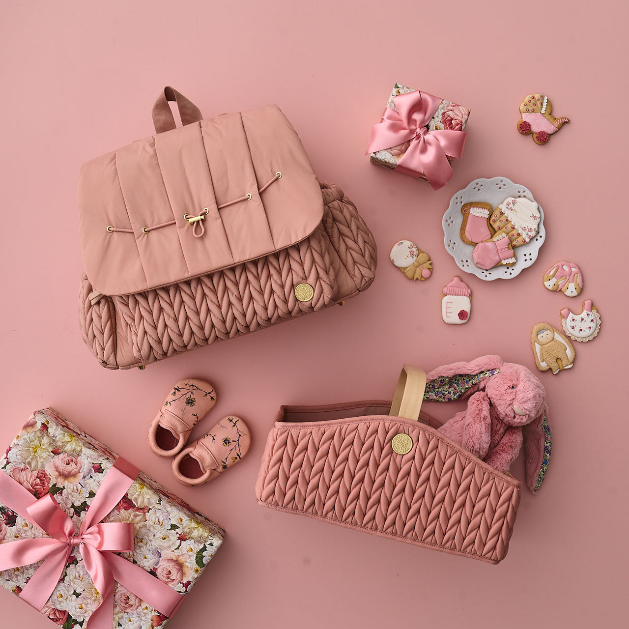 Flat lay with HAPP Levy Backpack and Diaper Caddy in matching rose, on a rose background with matching rose baby items, shown with two wrapped gifts in rose-pink theme.