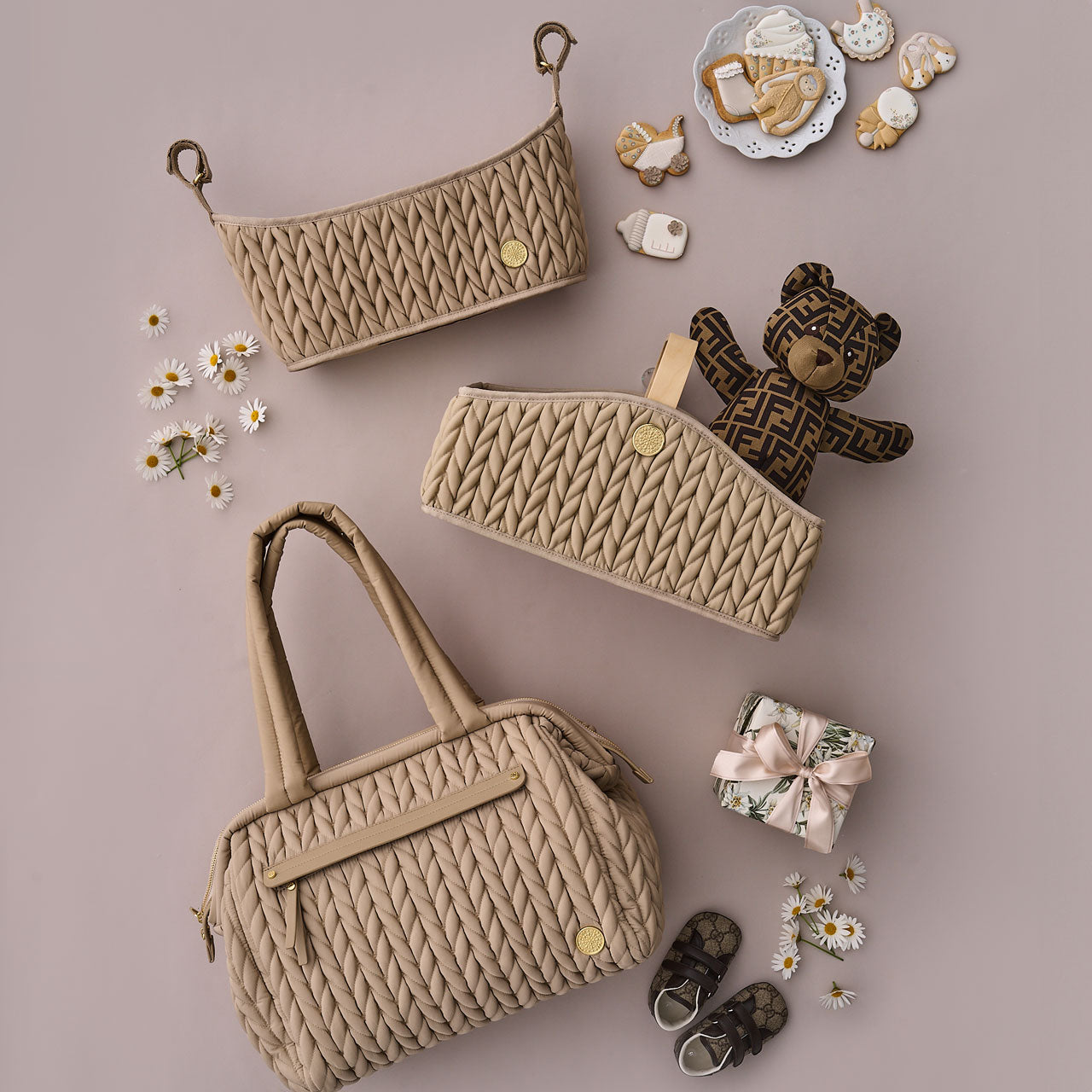 Paige Carryall Beige shown with HAPP Diaper Caddy and Stroller Organizer also in Beige, alongside white, beige, and brown baby items, a wrapped gift, and daisy flowers.