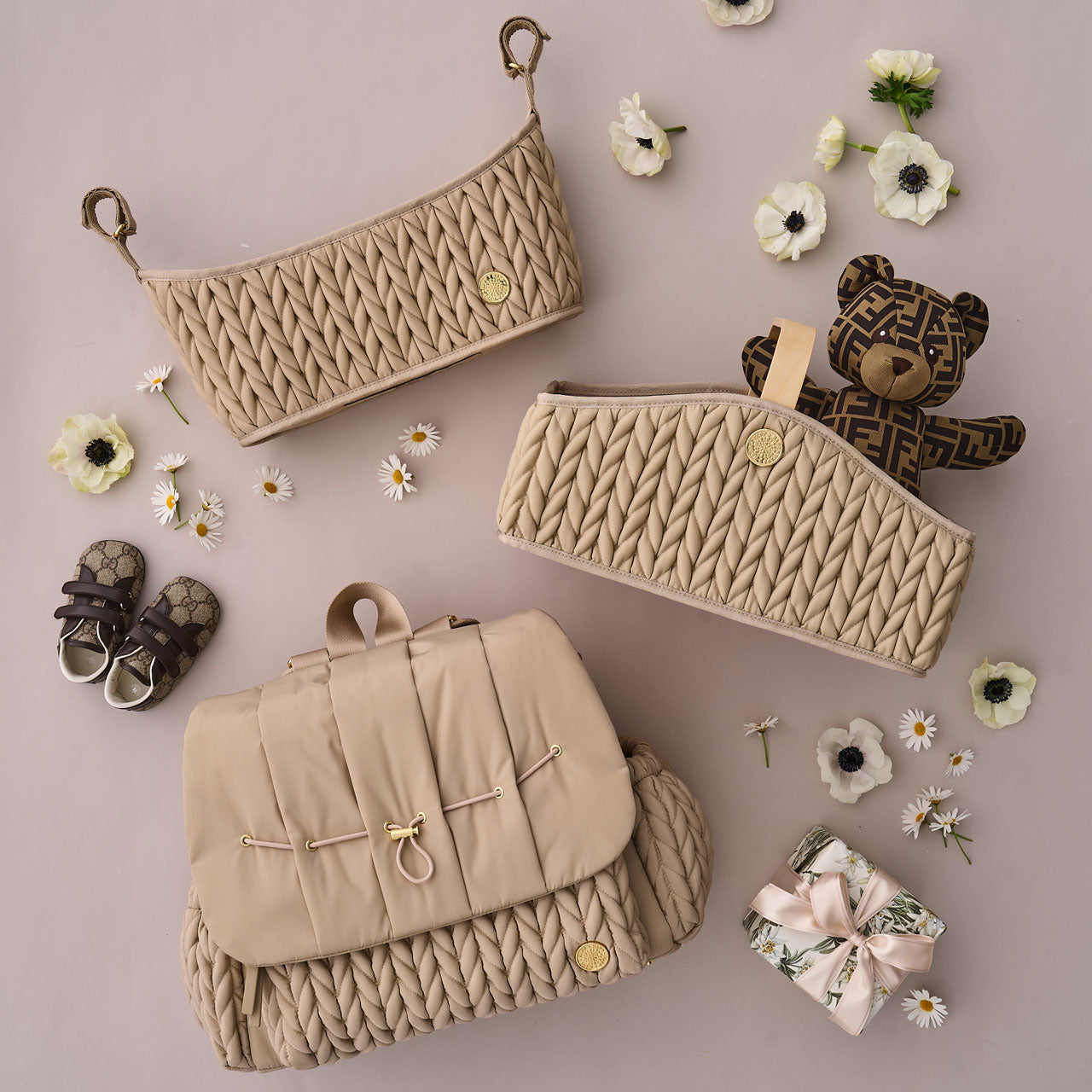Flat lay with HAPP Levy Backpack, Stroller Organizer, and Diaper Caddy, all in matching beige, arranged with baby items, a wrapped gift, and white flowers.