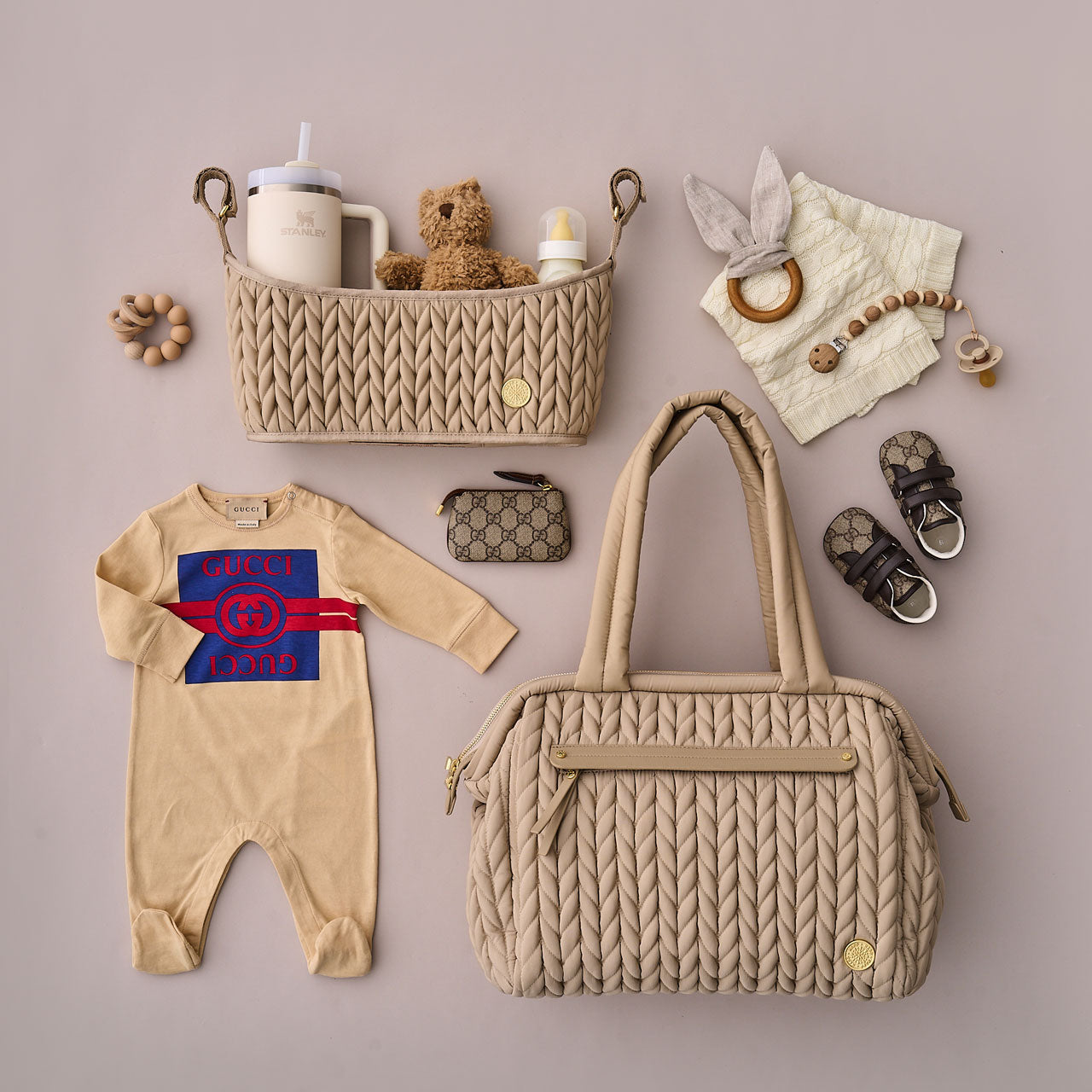 Paige Carryall Beige from the front with carry handles upright, next to beige and white baby and mom items.
