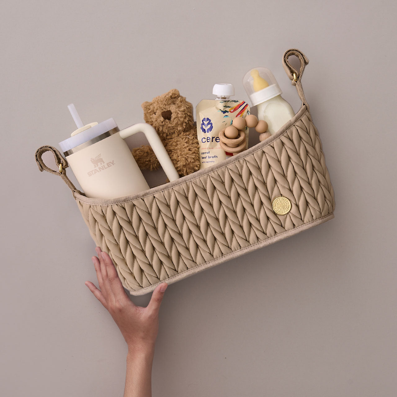 Beige stroller organizer packed with mom and baby items, held up by one hand to show the light weight of the caddy.