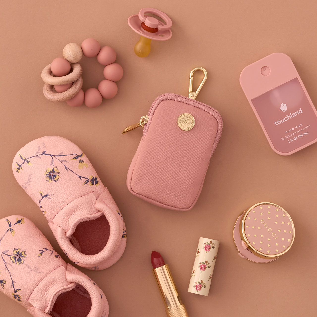 Rose Clip-on Pouch with gold clip, zipper, and logo, lying flat beside pink baby items on a pink background.