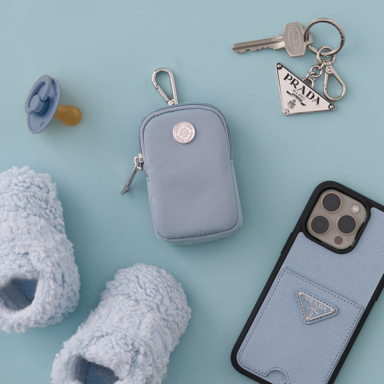 Blue Clip-on Pouch with silver clip, zipper, and logo, lying flat beside blue baby items on a blue background.