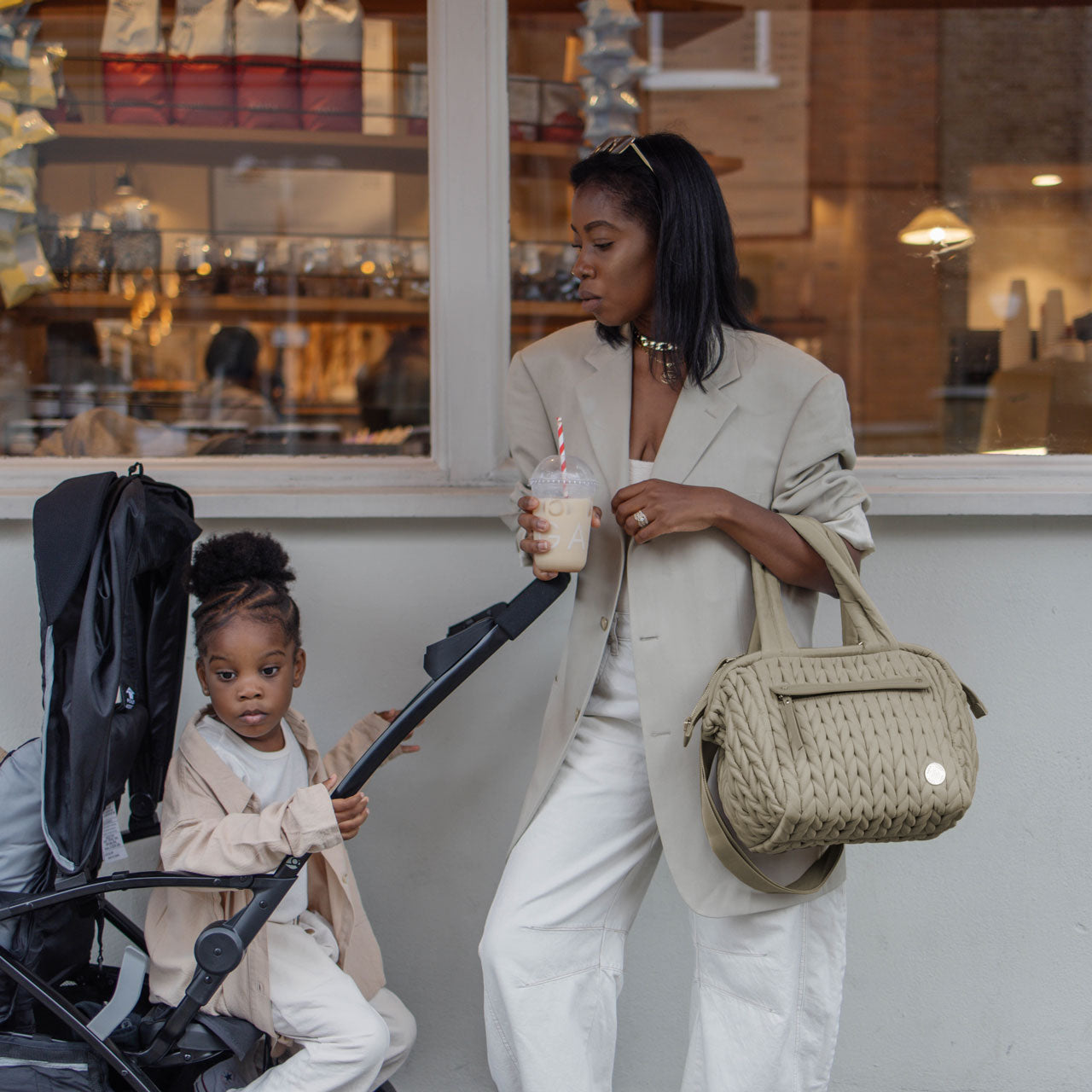 A mom carrying the Paige Mini Beige while walking with her toddler in a stroller, drinking a coffee and wearing a matching outfit with her bag and her daughter.