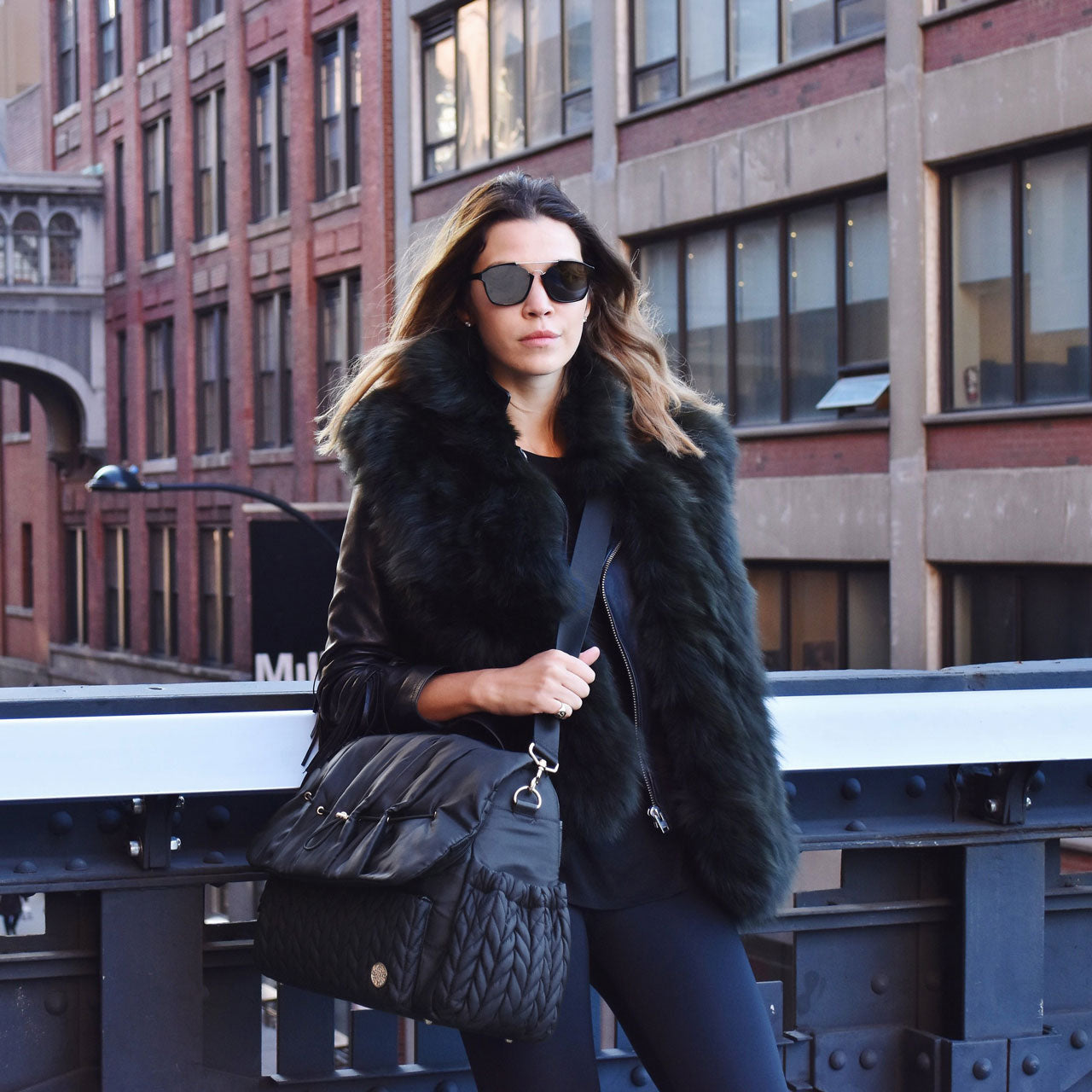 A stylish mom in all black and sunglasses stands on a bridge in NYC wearing her Levy Backpack Black over her shoulder by the detachable messenger strap, showing the versatility in carrying a HAPP diaper bag.