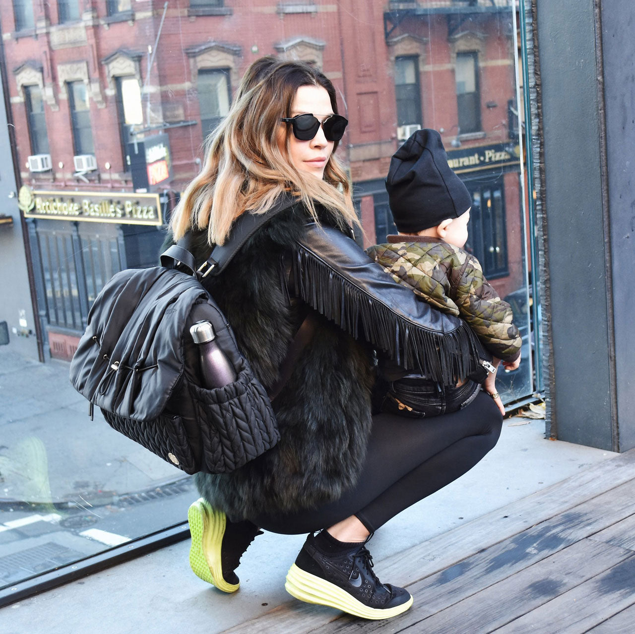 A NYC mom out and about with her son, her Levy Backpack Black on her back and her son in her arms, crouching down and looking over her shoulder at the camera.