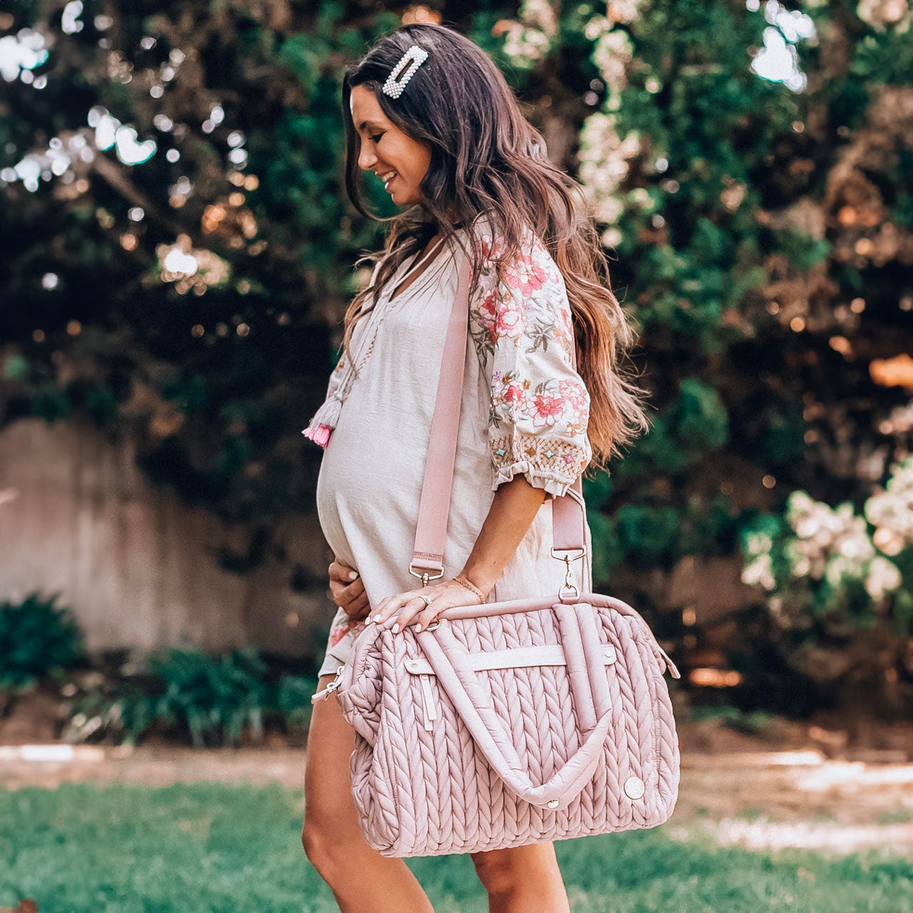 Pregnant mom in a sundress walking through a field carrying her Paige Carryall Dusty Rose by the detachable messenger strap.