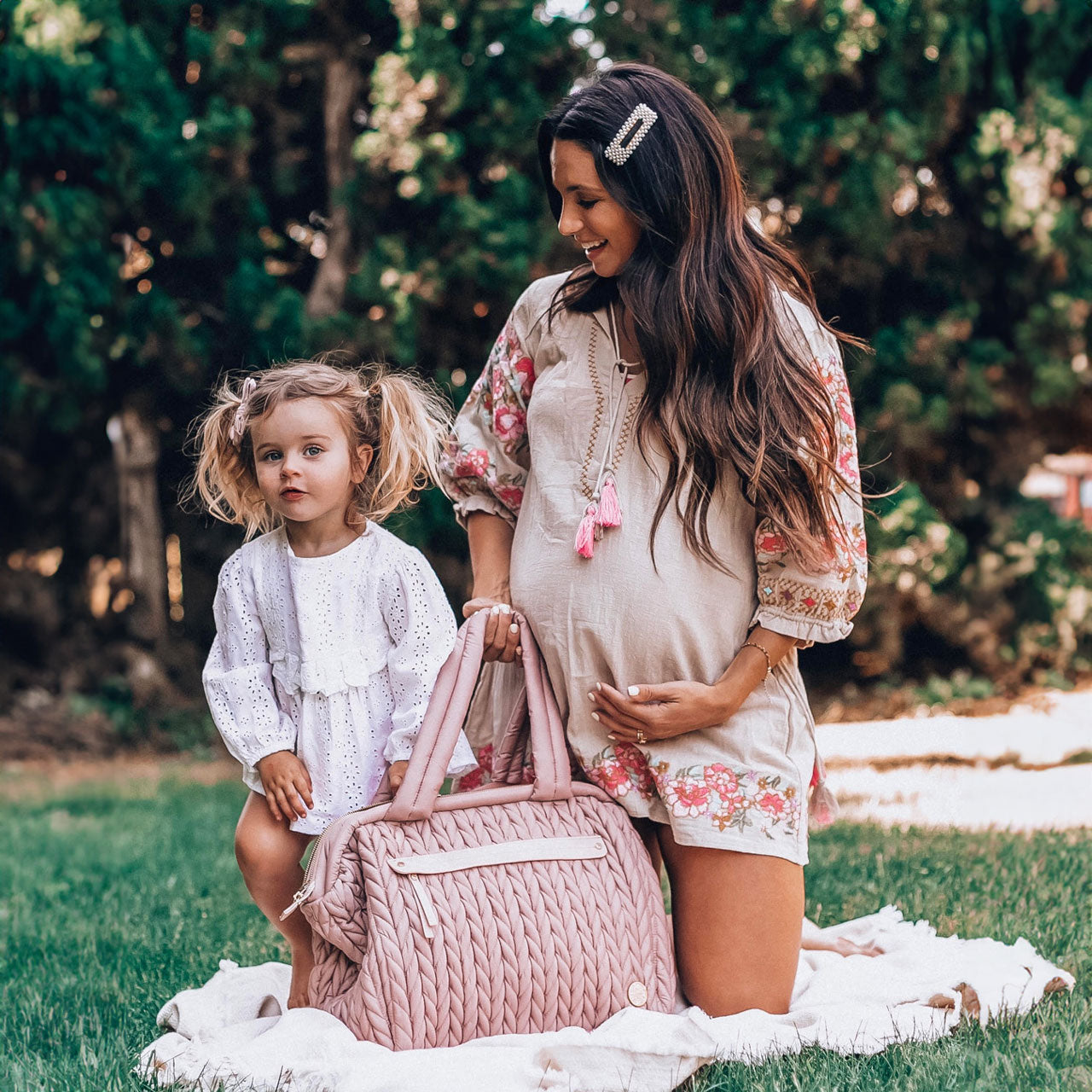 Pregnant mom with her toddler, both wearing sundresses, having a picnic in a field with their HAPP Paige Carryall in Dusty Rose.