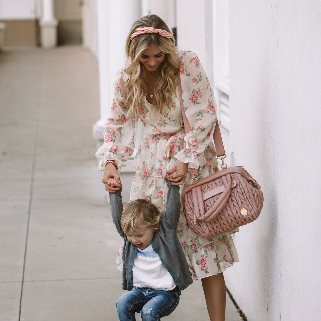 Mom carrying a Paige Mini Rose with the messenger strap over her shoulder, while holding her toddler's hands.