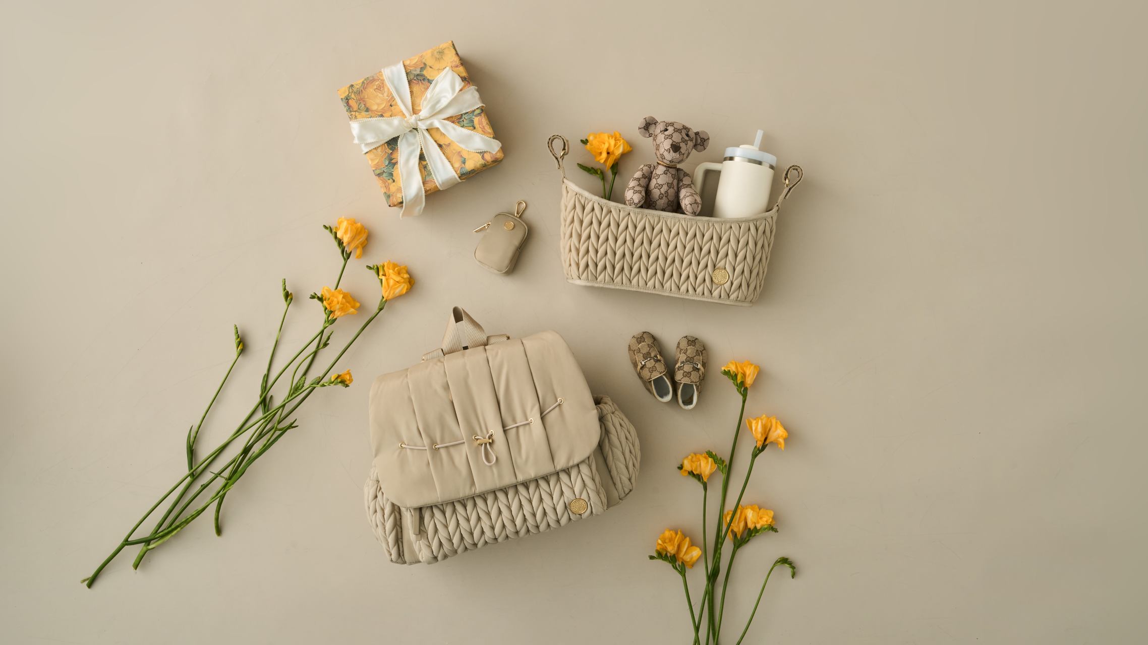 HAPP Brand Levy Backpack and Stroller Organizer in Signature Herringbone Beige pictured with Beige Clip-on Pouch, matching baby items, a wrapped yellow gift, and yellow flowers on a beige background.