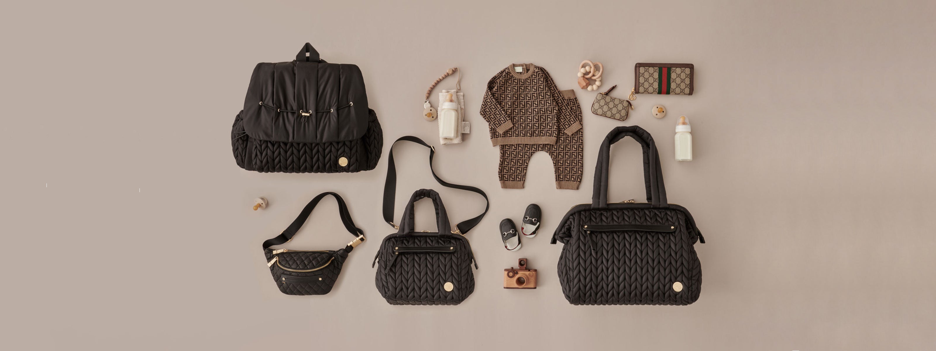 Levy Backpack, Paige Carryall, and Paige Mini, all in black, in a flat-lay photo with color-coordinated baby items on a beige background.
