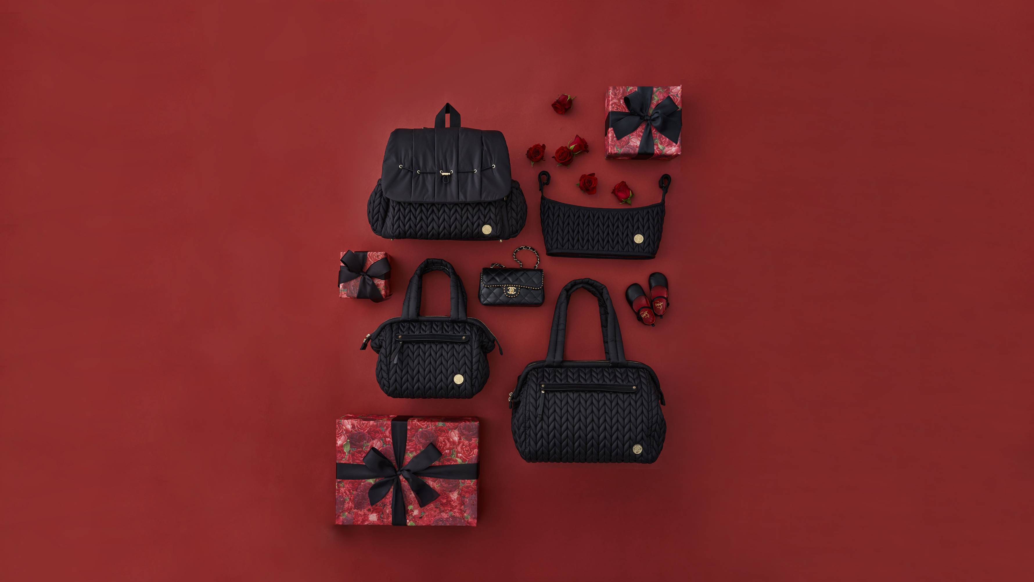 HAPP Brand Black Herringbone Collection with Levy Backpack, Paige Carryall, Paige Mini, and Stroller Organizer laid flat on a red background with black and red Valentine's gifts and accessories and red roses.