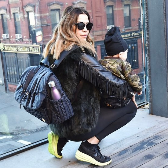 Stylish NYC mom wearing all black carrying Levy Backpack Black with padded shoulder straps, crouching with her son on her lap, looking over her shoulder at the camera.