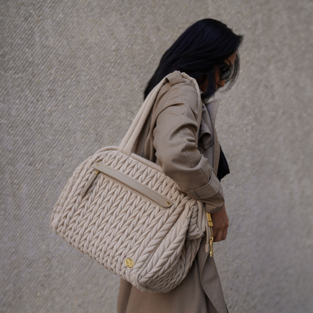 A fashionable mom walking with her Paige Carryall Beige over her shoulder by the padded handles, showing the quilted herringbone texture and gold hardware in natural light.