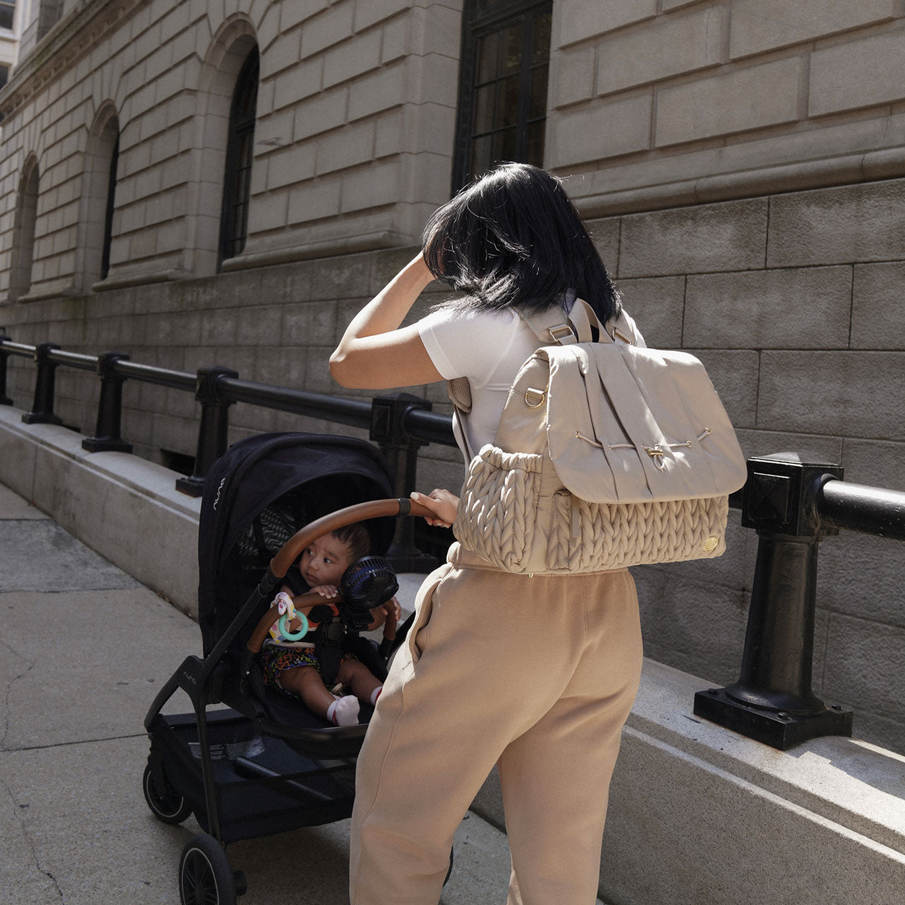 A stylish mom in beige and white walks down the sidewalk with her Levy Backpack Beige on her back, pushing her son in a stroller. Shows the luxe 3D quilted fabric and lustrous gold details.