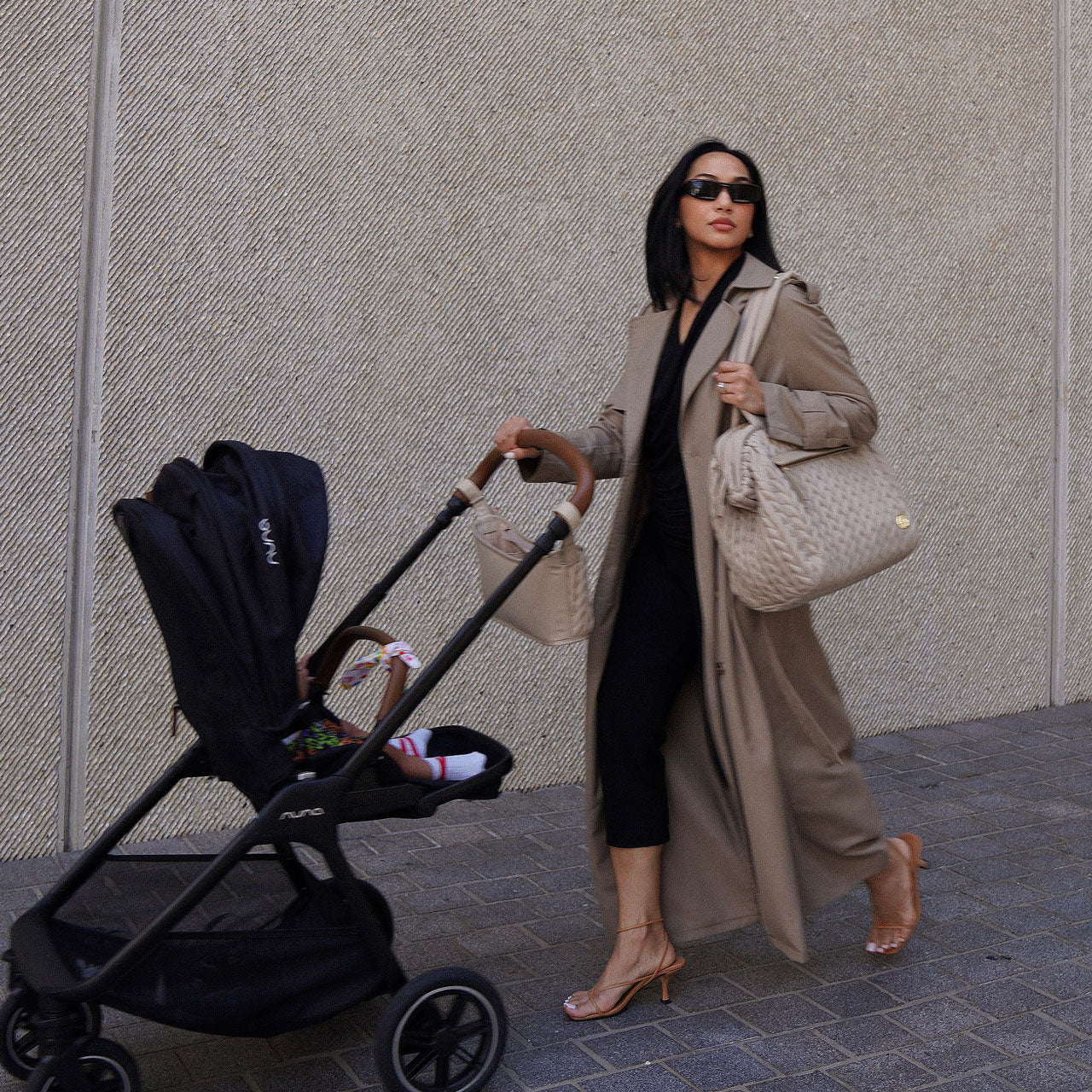 A mom pushes her baby in a stroller, with HAPP Stroller Organizer and Paige Carryall both matching in Beige.