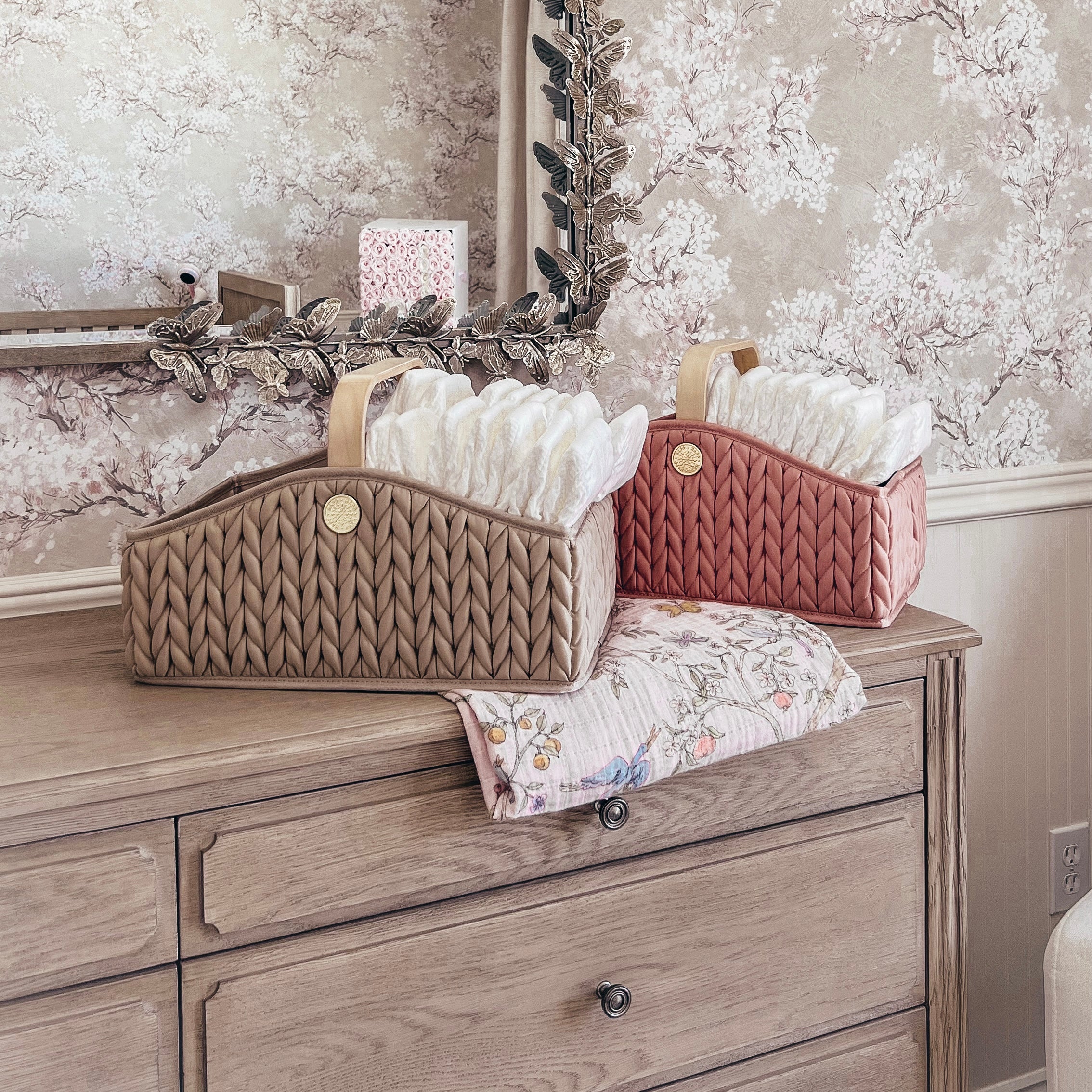 Pair of packed diaper caddies, Beige and Rose, sitting on the dresser in front of a mirror in a neutral-themed, floral-wallpaper nursery.
