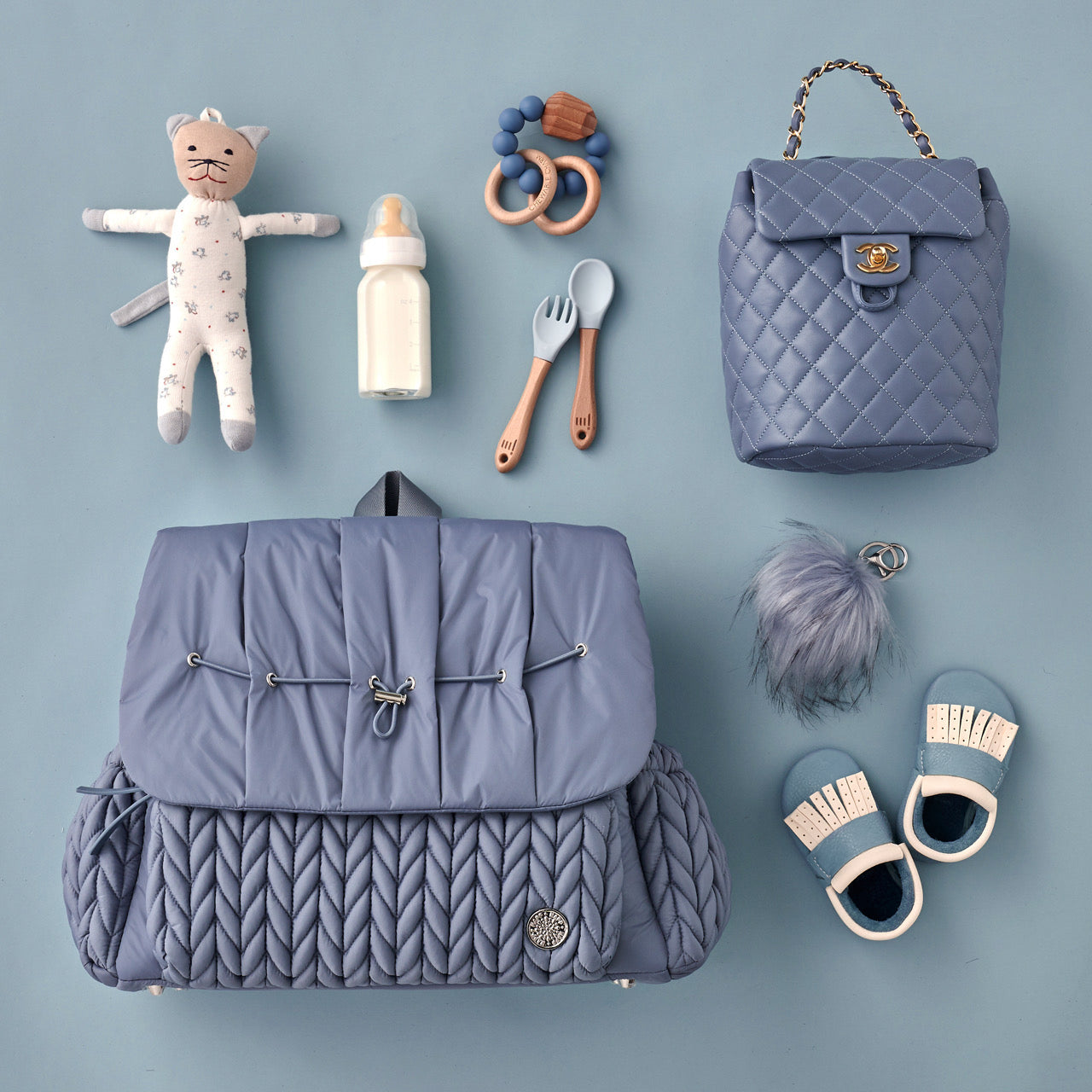 Levy Backpack in Ash Blue lying flat, showing the luxe quilted fabric, silver hardware details, convenient exterior bottle pockets, and top carry handle. Arranged with white and blue baby items on a blue background.