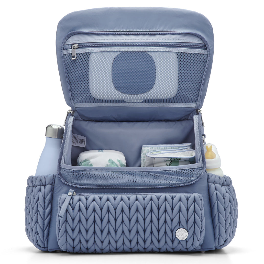 Levy Backpack Ash Blue standing upright with the flap open to show the interior mesh pocket and 4 interior pockets. Bottles in the exterior bottle pockets. The bag is lined with Blue fabric.