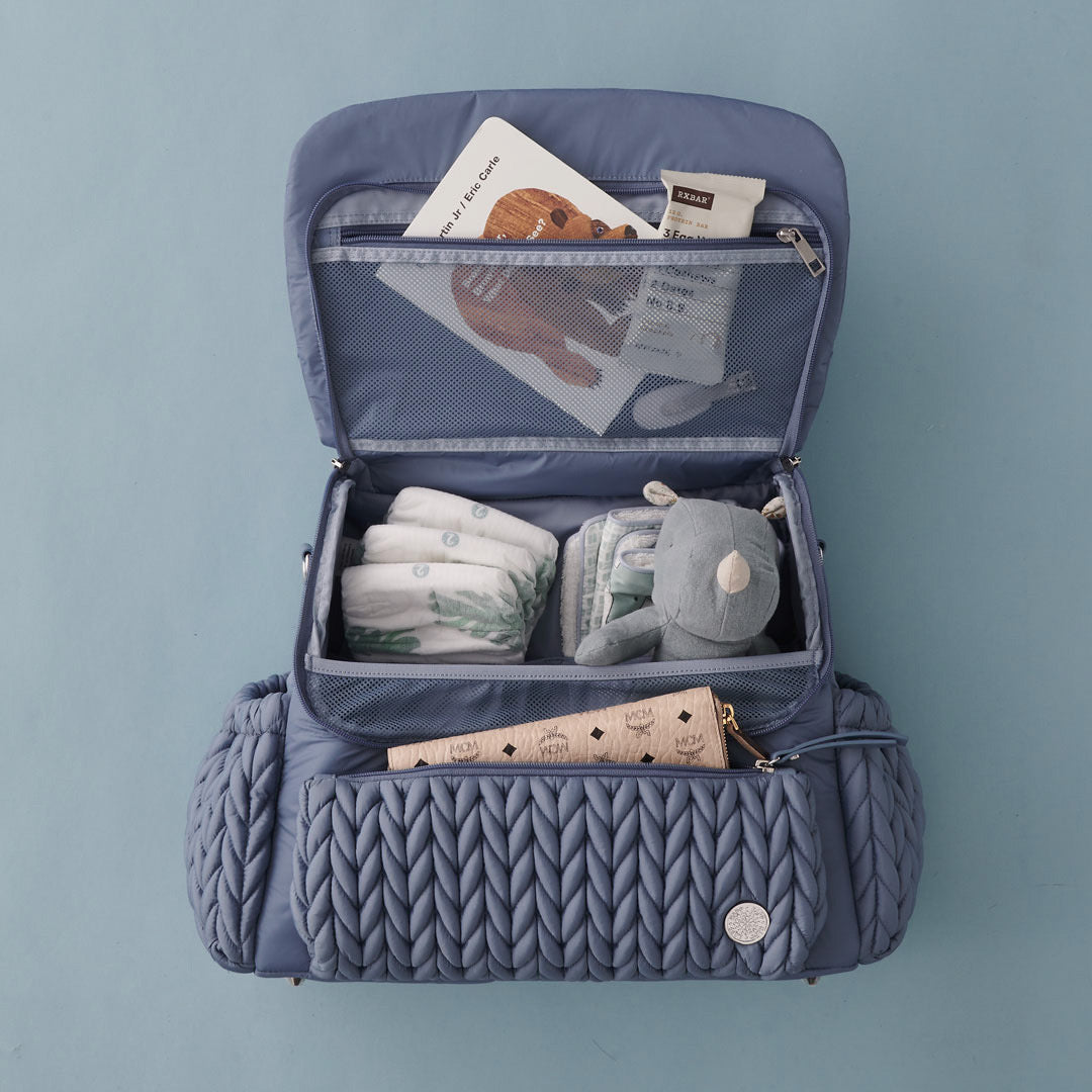 Levy Backpack Ash Blue lying flat, with top flap open, packed with white and blue baby items. Shows front mommy pocket, interior mesh pocket, and superior organization of 4 interior pockets.