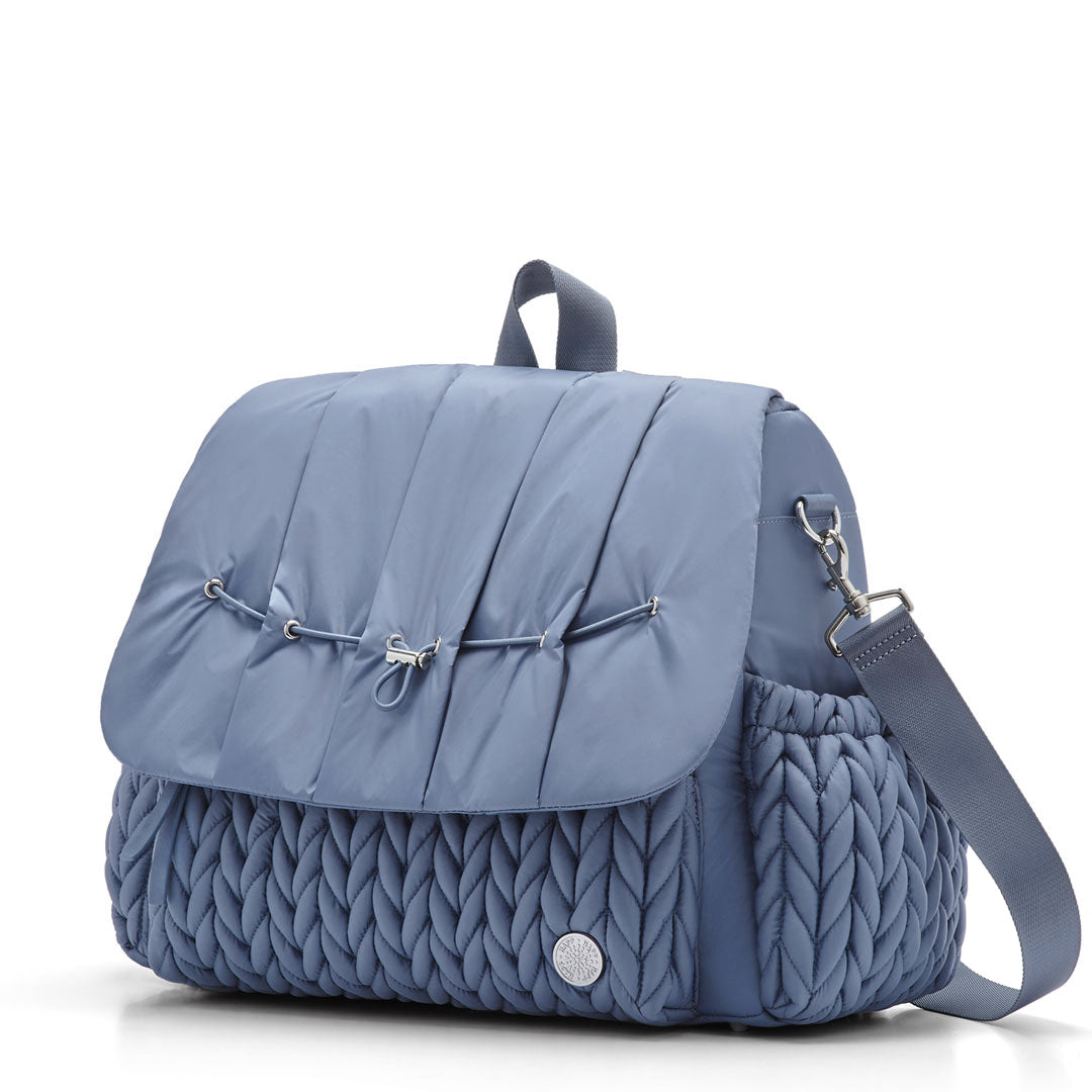 3/4 view of Levy Backpack Ash Blue, standing upright and zipped closed, with detachable messenger strap attached. Shows luxe, 3D herringbone fabric, top carry strap, and silver hardware details.