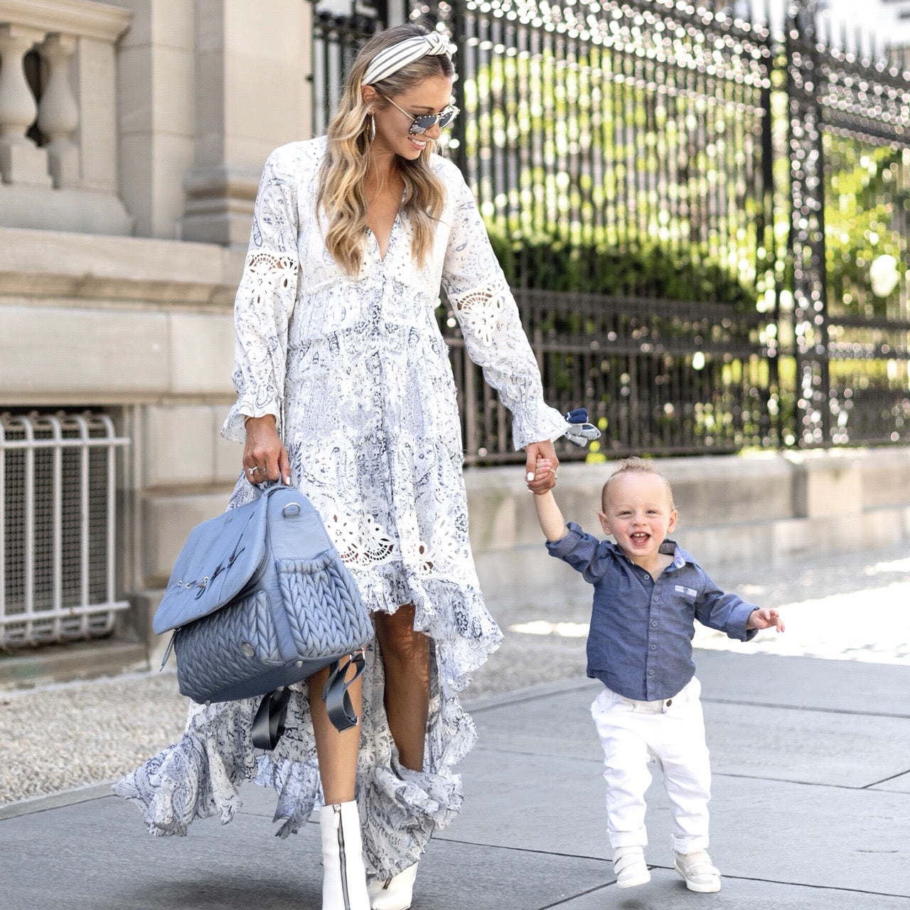 A mom in a white and blue sundress walks down the sidewalk holding her toddler son's hand in one hand and her Levy Backpack Ash Blue by the carry handle in the other. Shows how you can dress a Levy up or down.