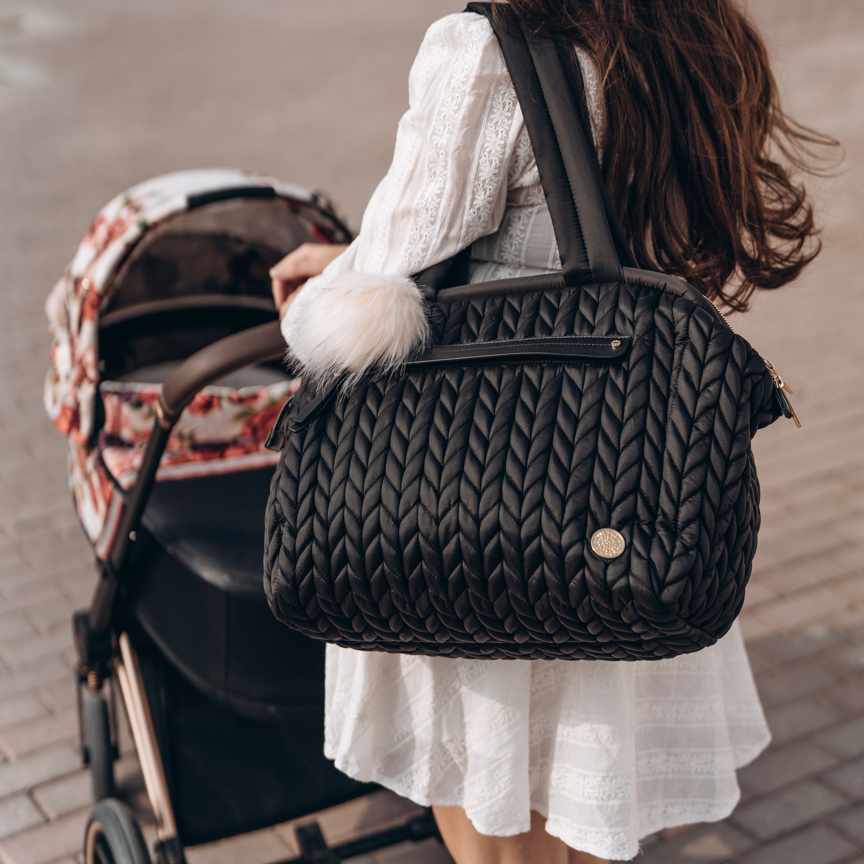 Chic mom in white dress carrying Paige Carryall Black over her shoulder by the padded handles, pushing a pink stroller with floral pattern on brick walkway.