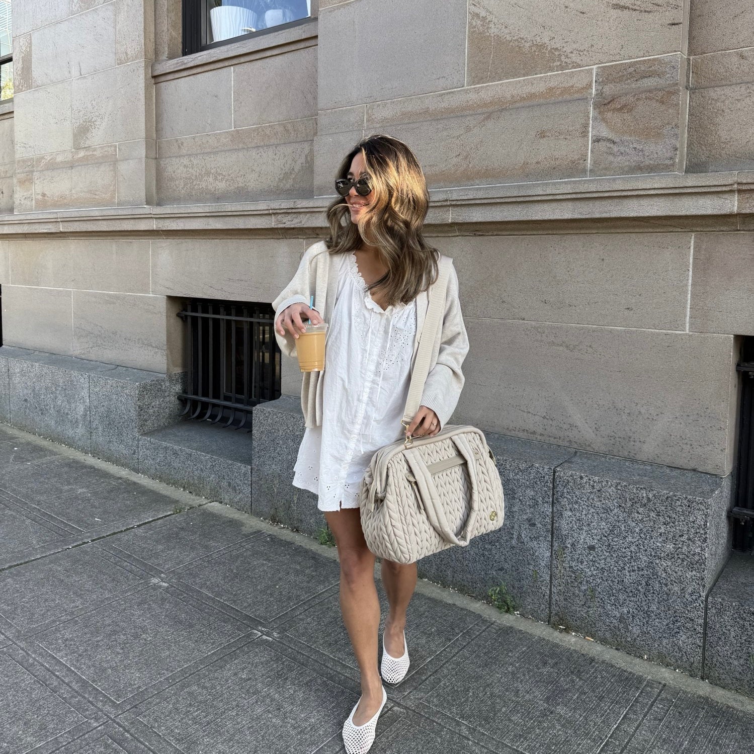A stylish mom in a white sundress and white flats carrying a HAPP Brand Paige Carryall Diaper Bag in Beige Herringbone, walking outside and carrying an iced coffee.