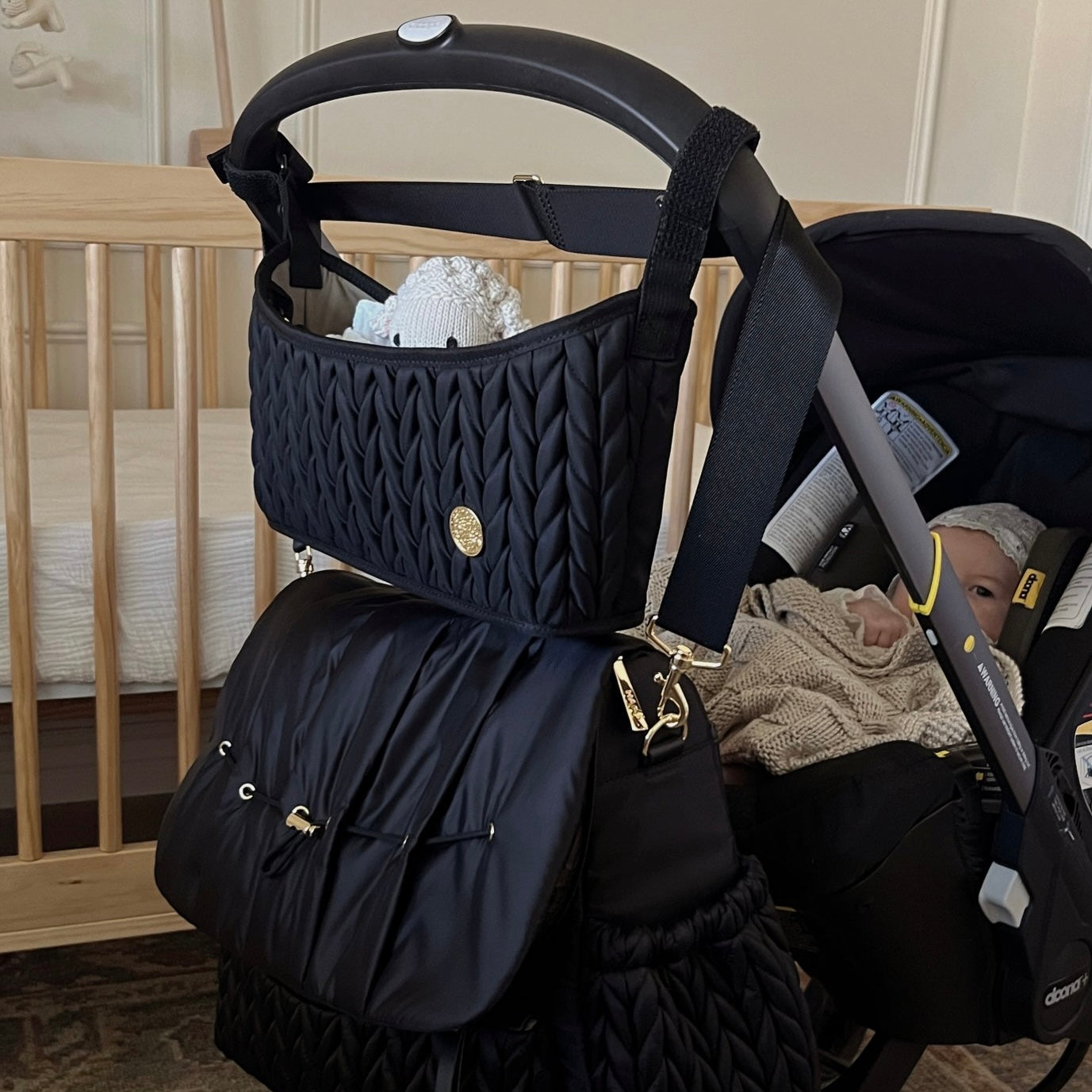 A stroller with a baby in front of a crib, with HAPP Brand Levy Backpack Diaper Bag and Stroller Organizer, both in Black Herringbone, attached to the handle of the matching black stroller.