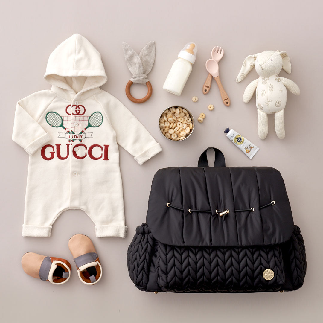 Levy Backpack in Black lying flat, showing the luxe quilted fabric, gold hardware details, convenient exterior bottle pockets, and top carry handle. Arranged with white and beige baby items on a beige background.