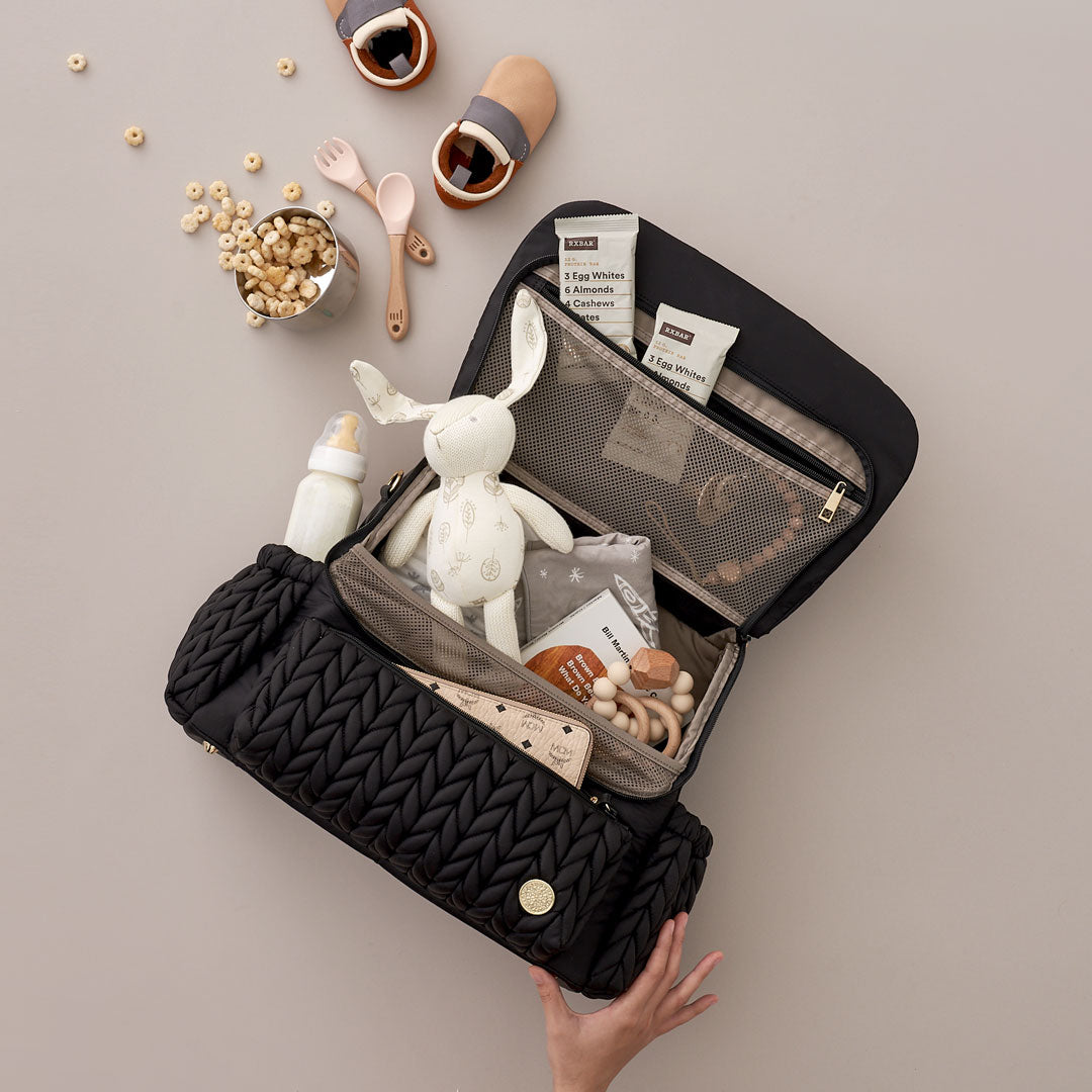 Levy Backpack Black lying flat, with top flap open, packed with white and beige baby items. Shows front mommy pocket, interior mesh pocket, and superior organization of 4 interior pockets. A hand holds up the bag from the bottom to demonstrate its light weight.