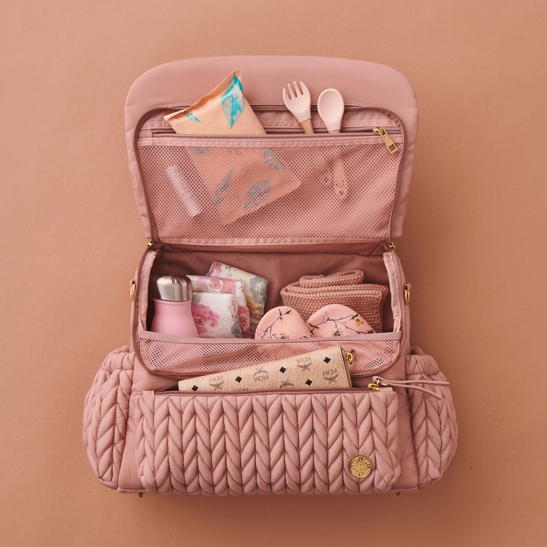 Levy Backpack Dusty Rose lying flat, with top flap open, packed with white and pink baby items. Shows front mommy pocket, interior mesh pocket, and superior organization of 4 interior pockets.